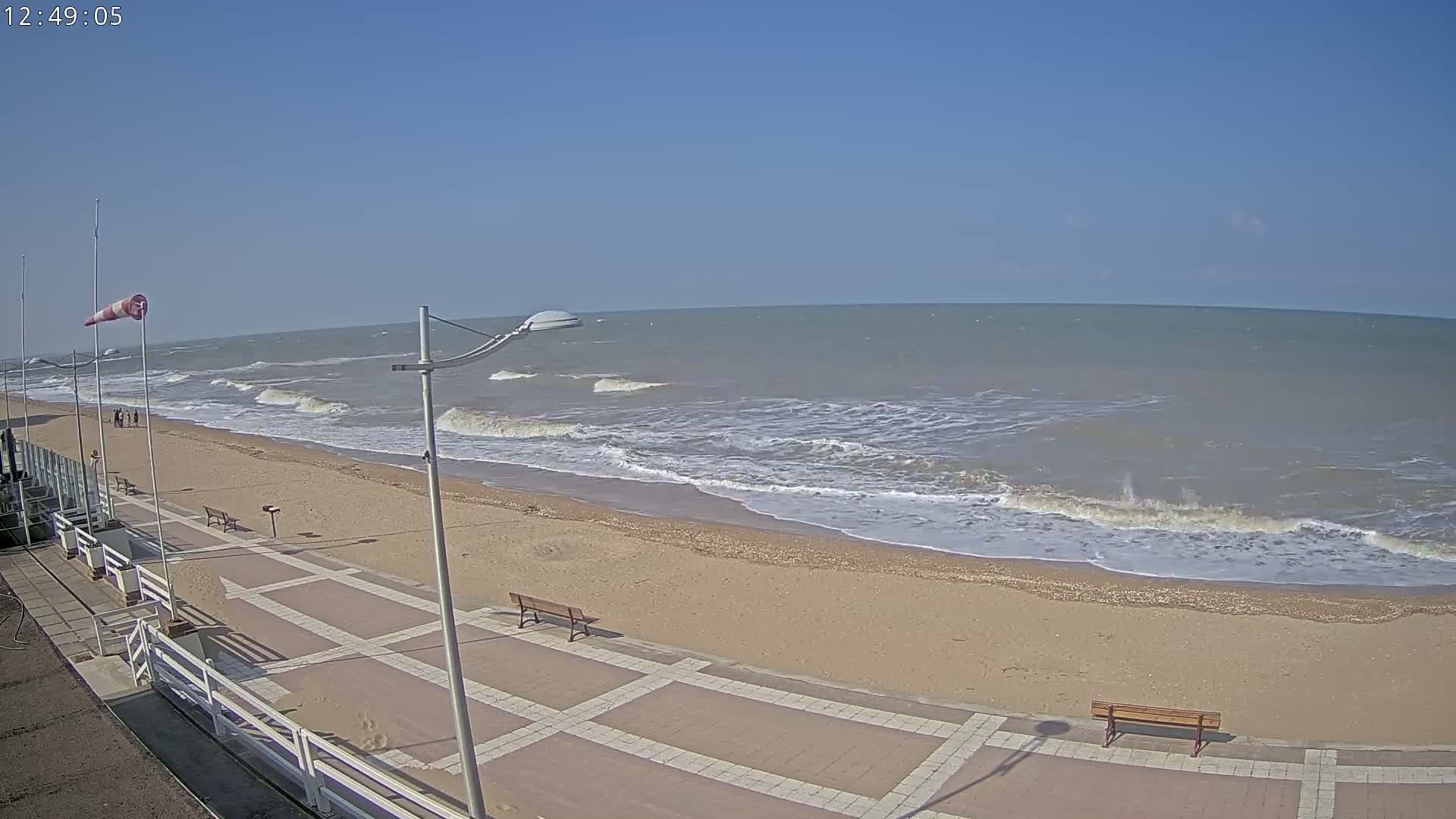 A sandy beach with a paved walkway and benches is shown, with moderate waves rolling onto the shore under a clear, sunny sky.