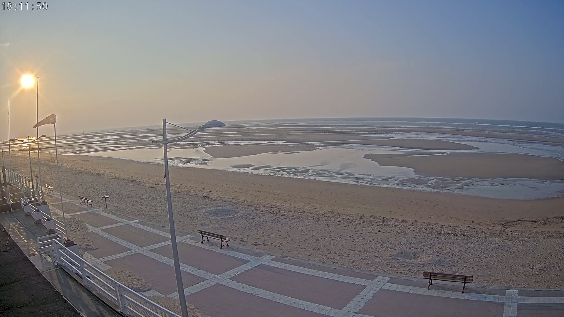 A sandy beach with shallow, receding water at low tide is seen from a high vantage point on a clear, sunny day.