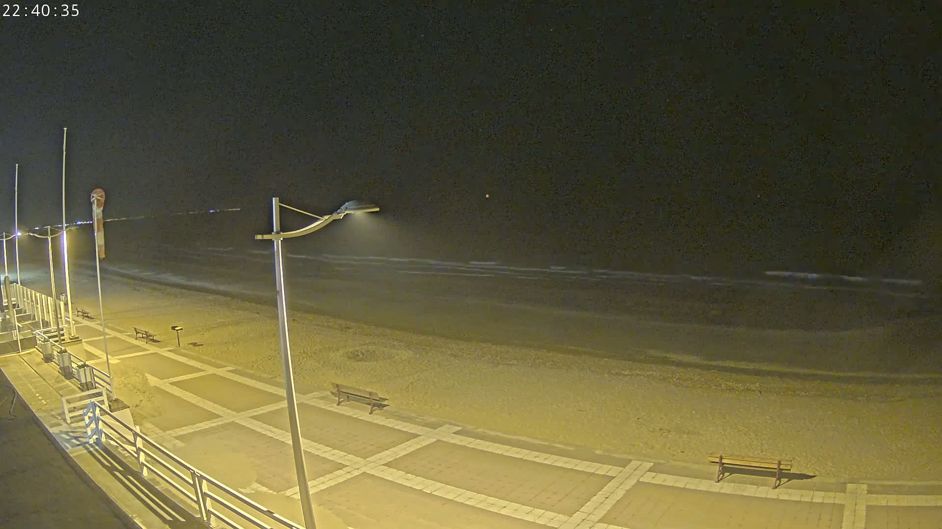A nighttime view of a sandy beach with gentle waves under a clear, dark sky, showing a paved walkway with benches and lampposts.