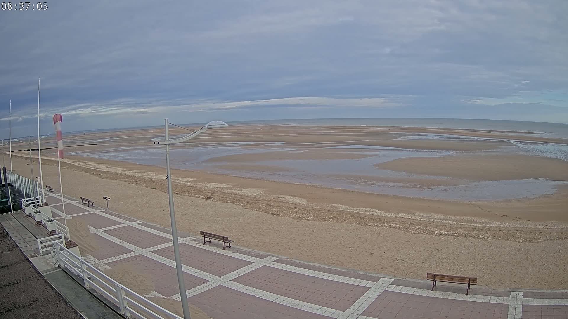 A paved walkway overlooks a wide, sandy beach with shallow pools of water under an overcast sky.