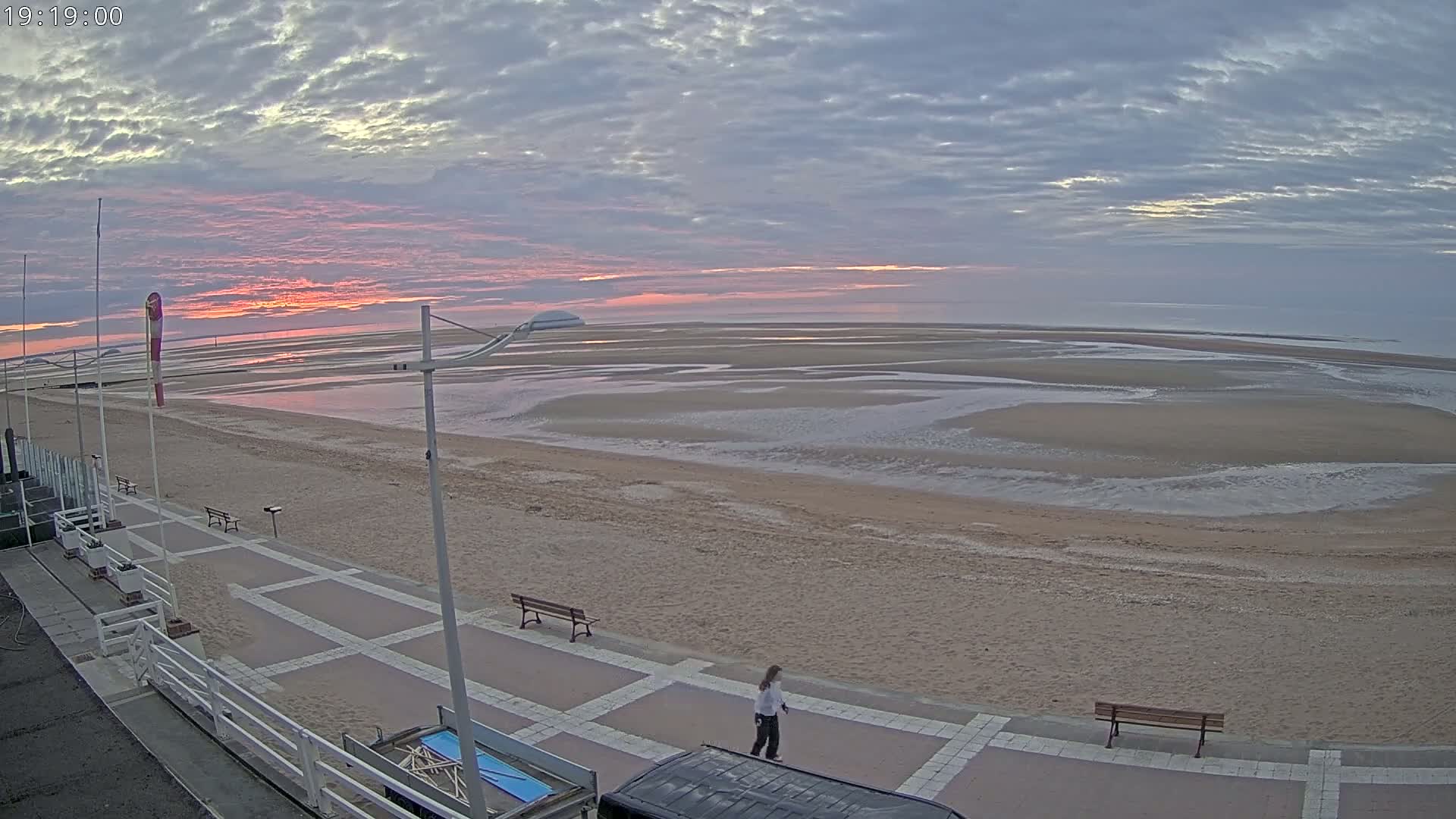 A person walks along a paved beachfront path overlooking a wide, sandy beach at low tide under a partly cloudy sky with a pink sunset.