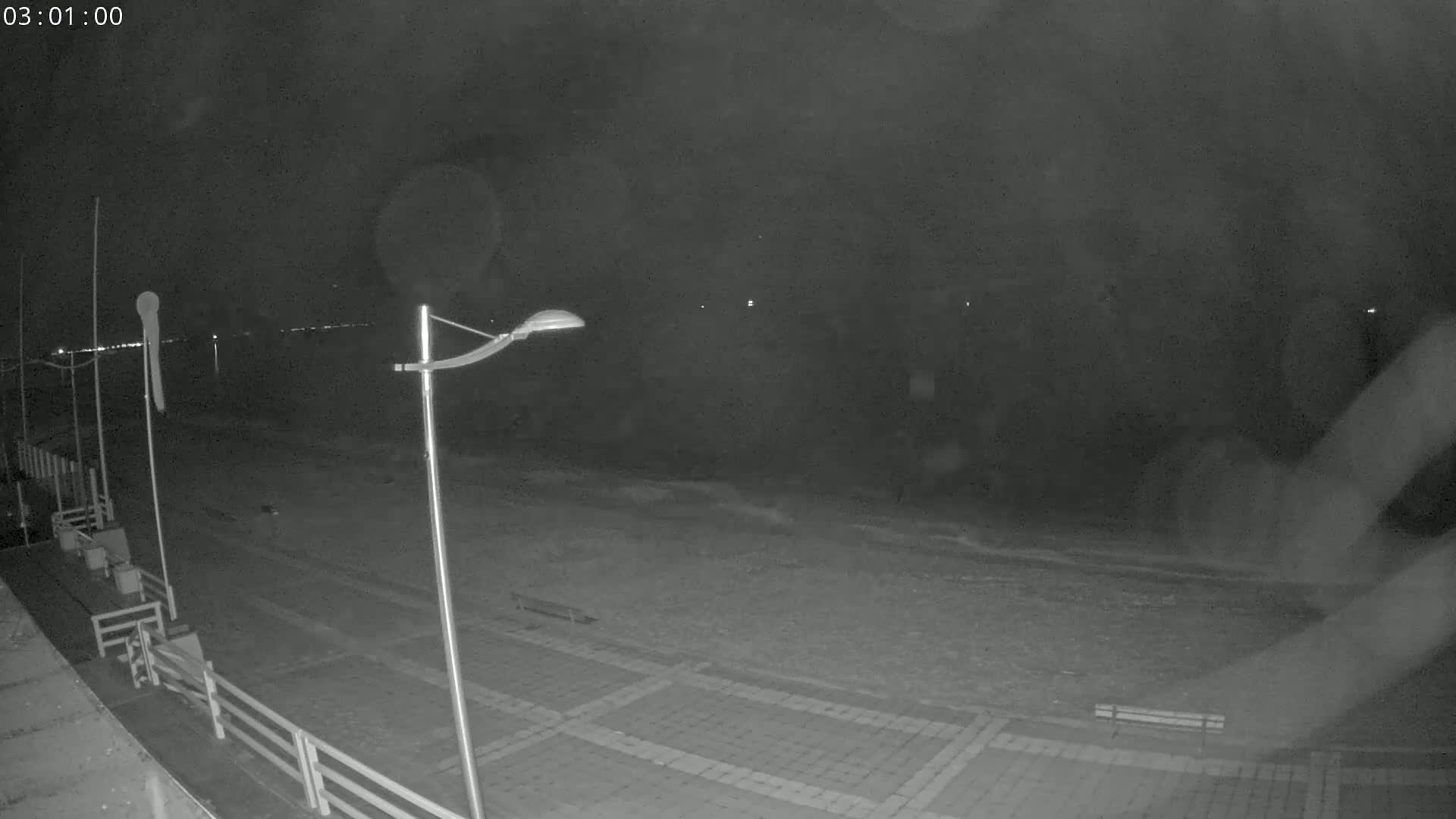 A nighttime, low-light view of a paved beachfront area with a lamppost, some benches, and a calm sea in the background.