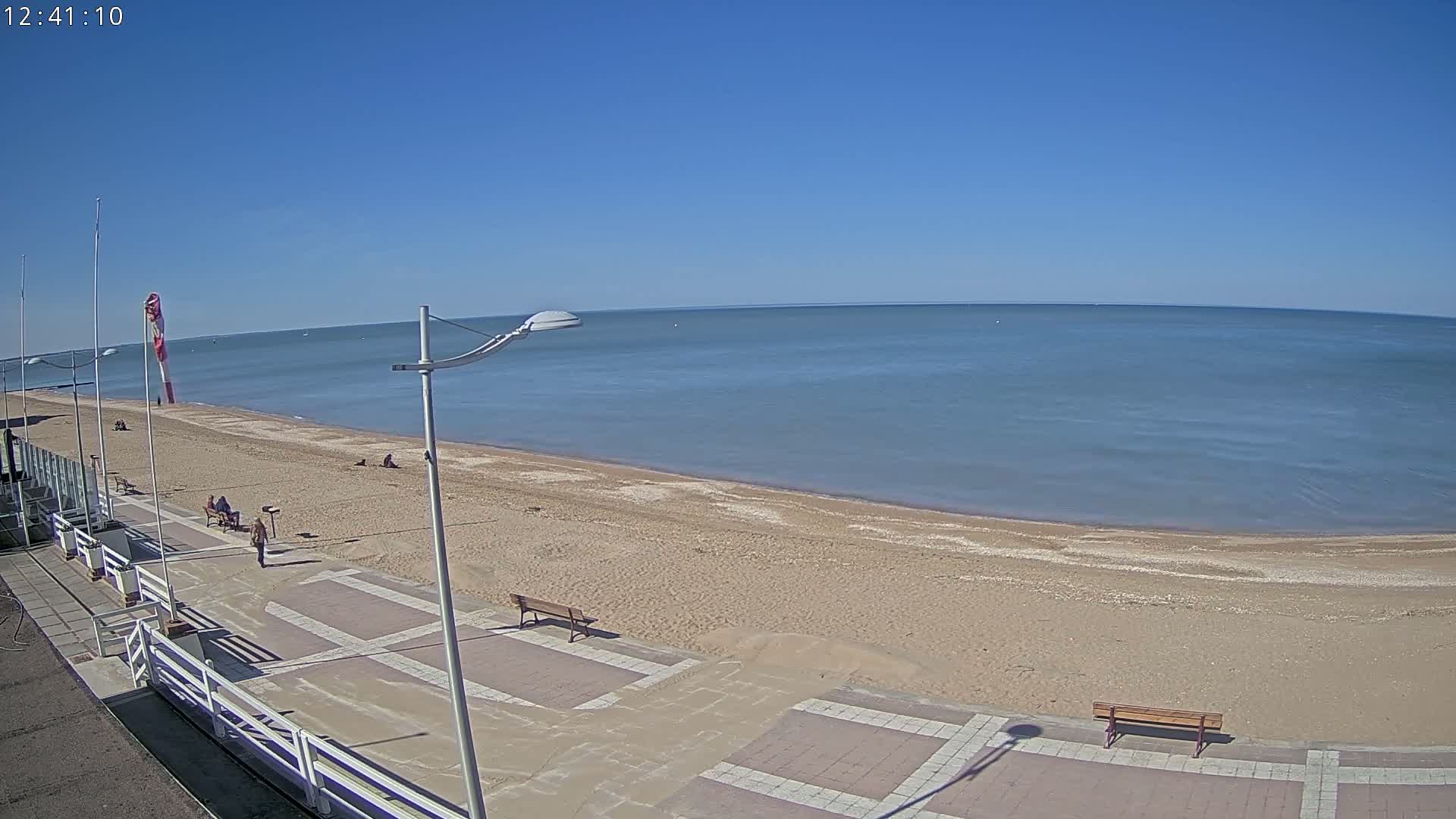 A sandy beach with a paved walkway and several people is visible, under a clear blue sky.