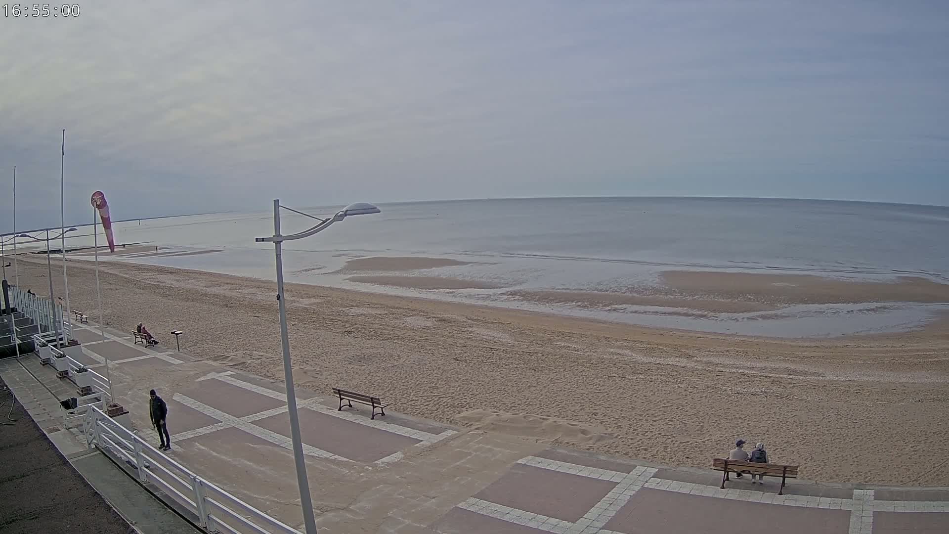 A wide shot of a sandy beach with a calm, overcast sea, featuring a paved promenade with several benches and a few people scattered along it.