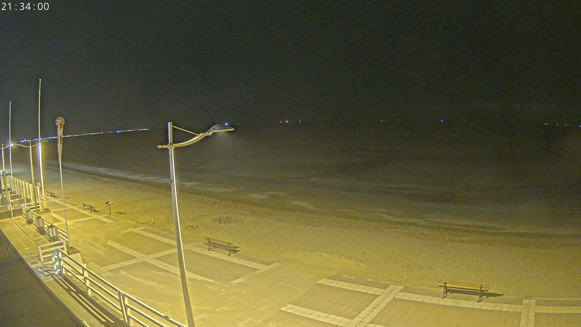 A nighttime view of a mostly empty beach with a paved walkway and benches, gently lapping waves, and distant lights.