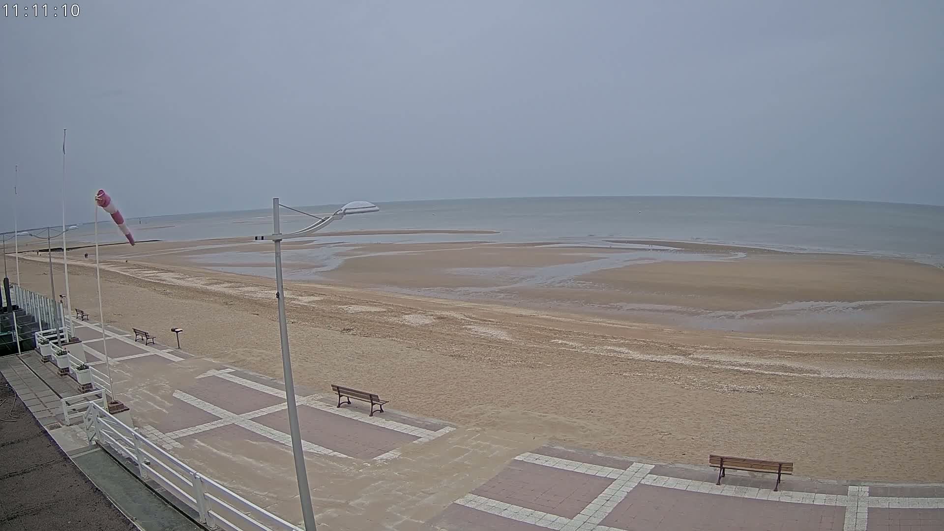 A mostly overcast day reveals a sandy beach with benches and a boardwalk overlooking a calm sea at low tide.