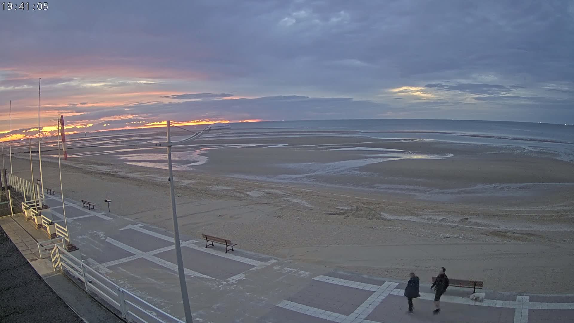 A wide, sandy beach at sunset with a paved walkway, benches, and two people walking a small dog.