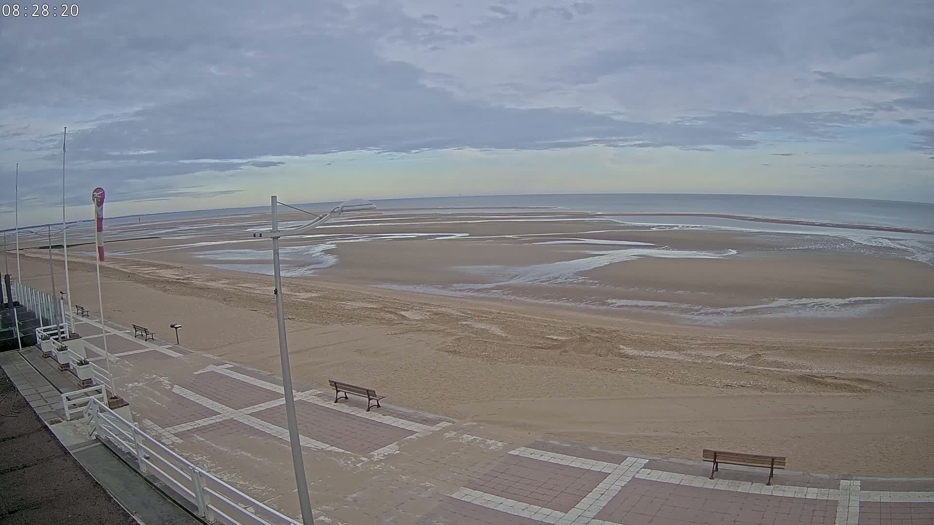 A wide sandy beach with scattered pools of water stretches to a calm ocean under a cloudy sky,  with a paved walkway and benches visible in the foreground.
