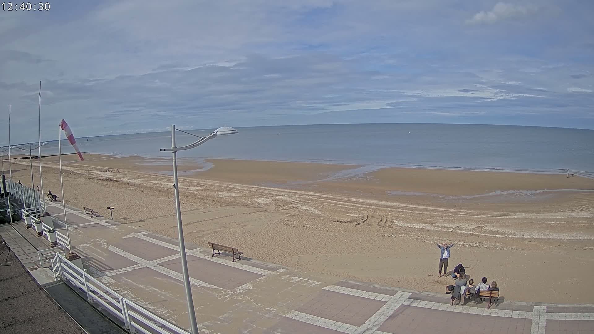 A mostly sunny day shows a wide sandy beach with a few people on it and several more seated on benches along a paved walkway next to the water.