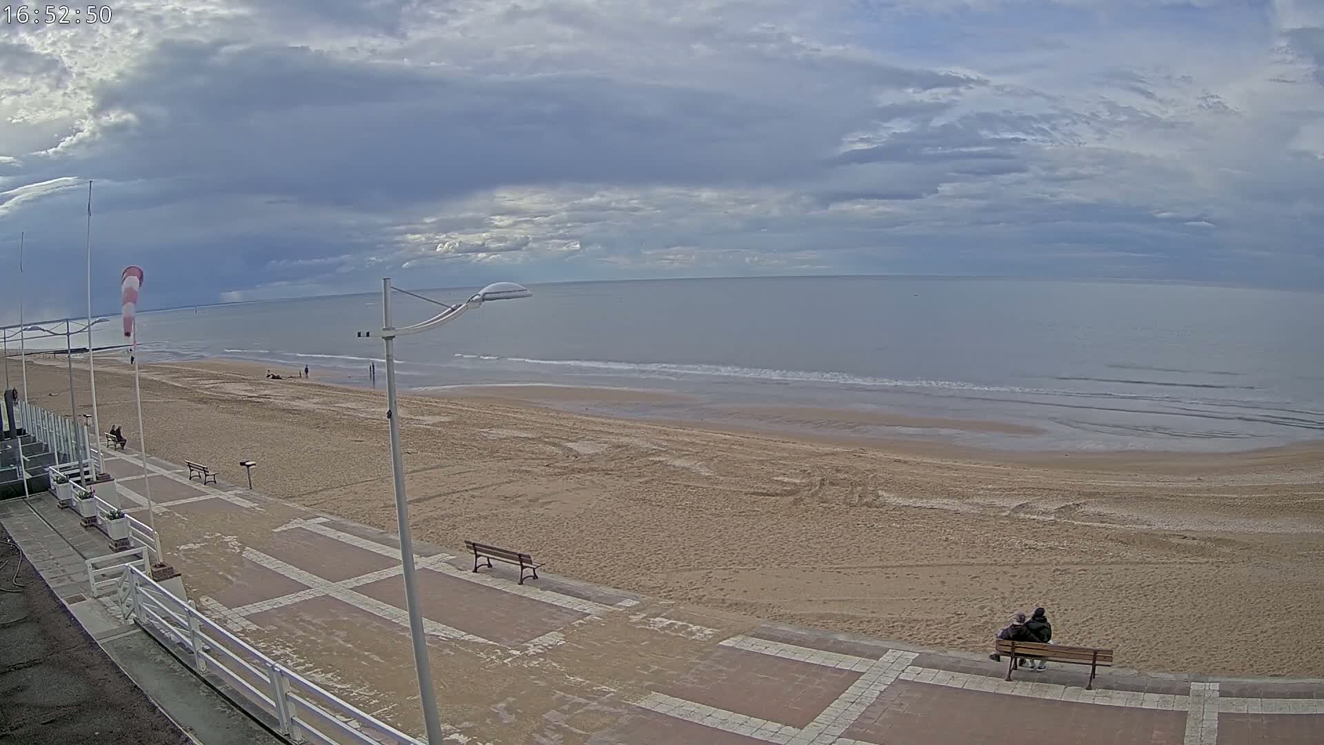 A mostly cloudy day shows a wide sandy beach with a calm ocean, a paved walkway, benches, and a couple sitting on one of the benches.