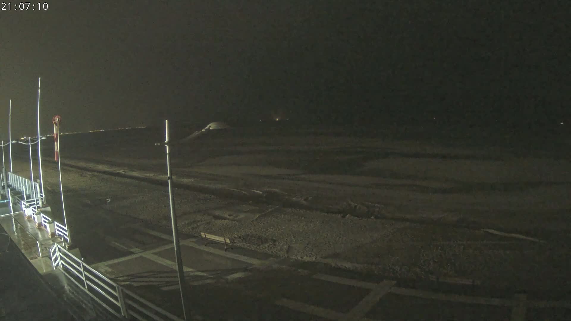 A nighttime view of a dark, wet beach with a few lights visible in the distance under a cloudy sky.