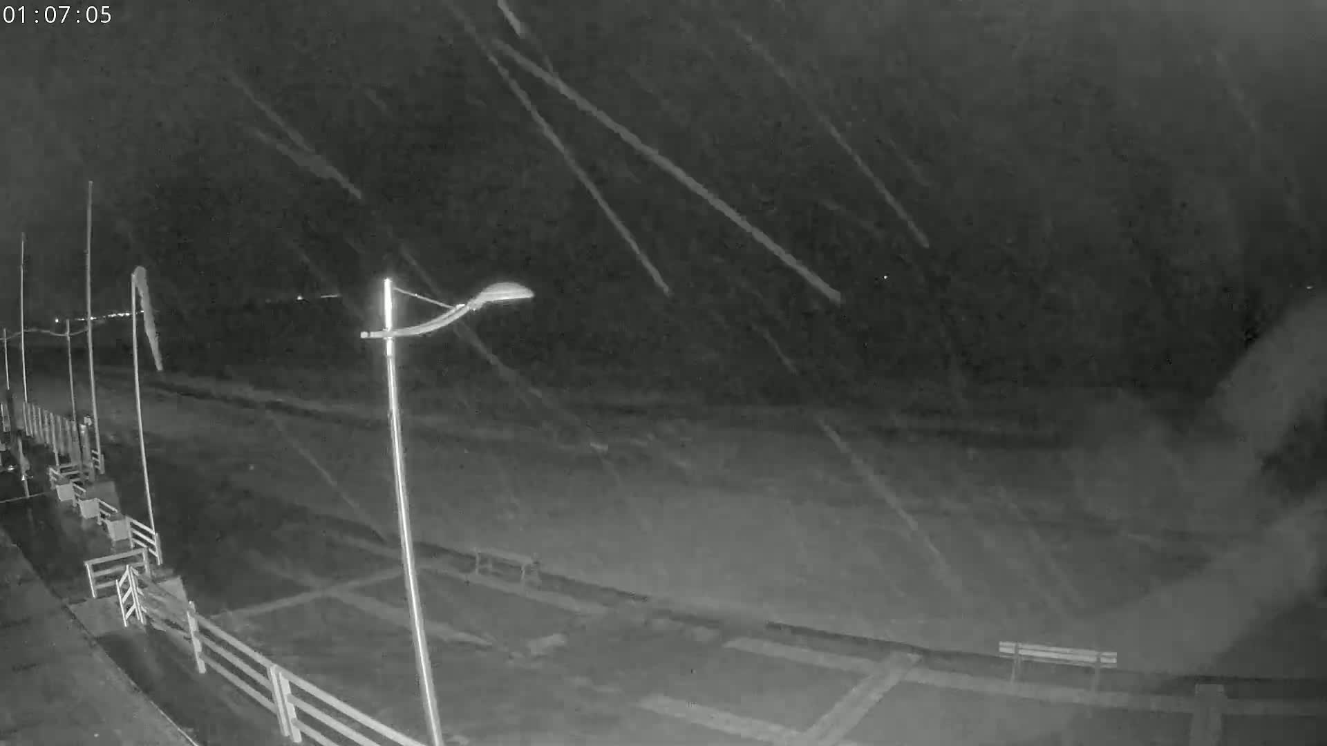 A nighttime, snowy scene shows a mostly empty pier or boardwalk with lights and railings under a heavy snowfall.