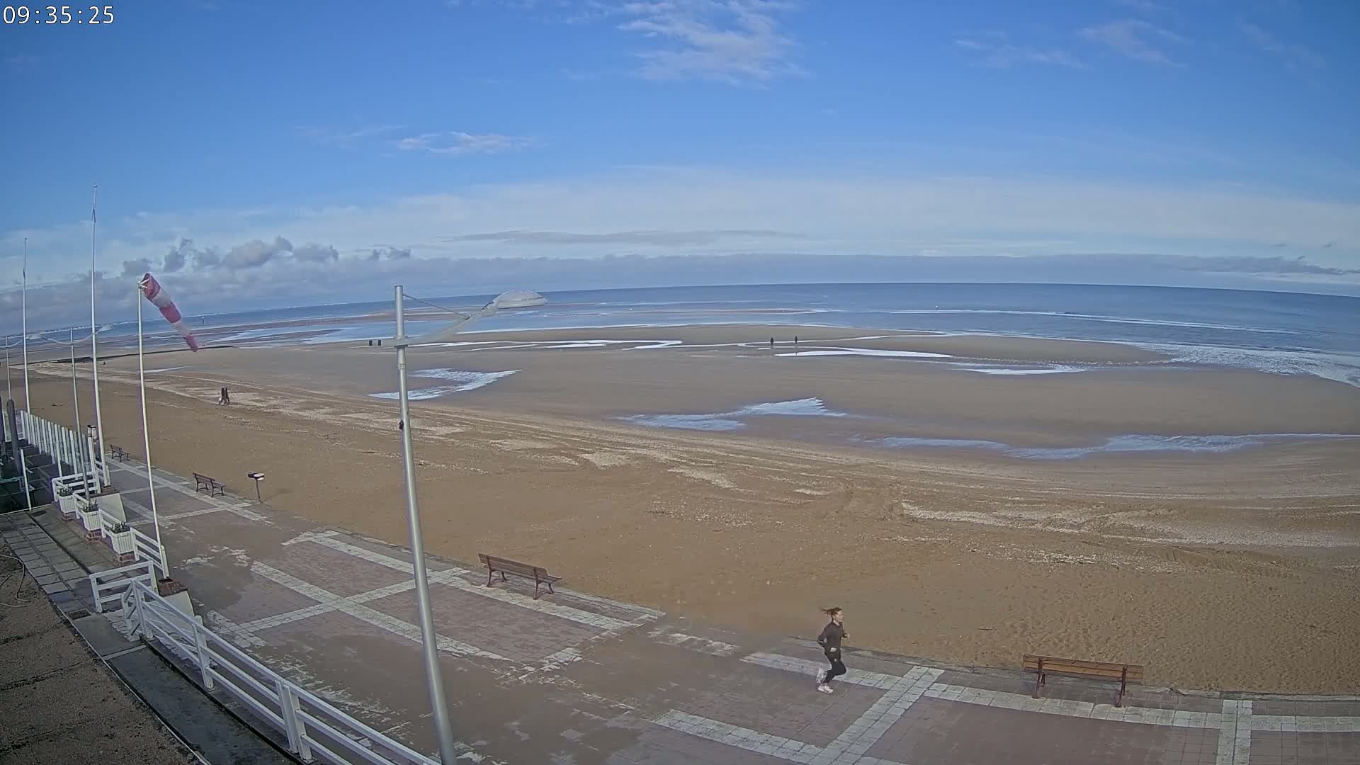 A mostly sunny day at a beach shows a wide expanse of sand with patches of water, a person jogging along a paved walkway near the water's edge, and a few people walking on the beach in the distance.