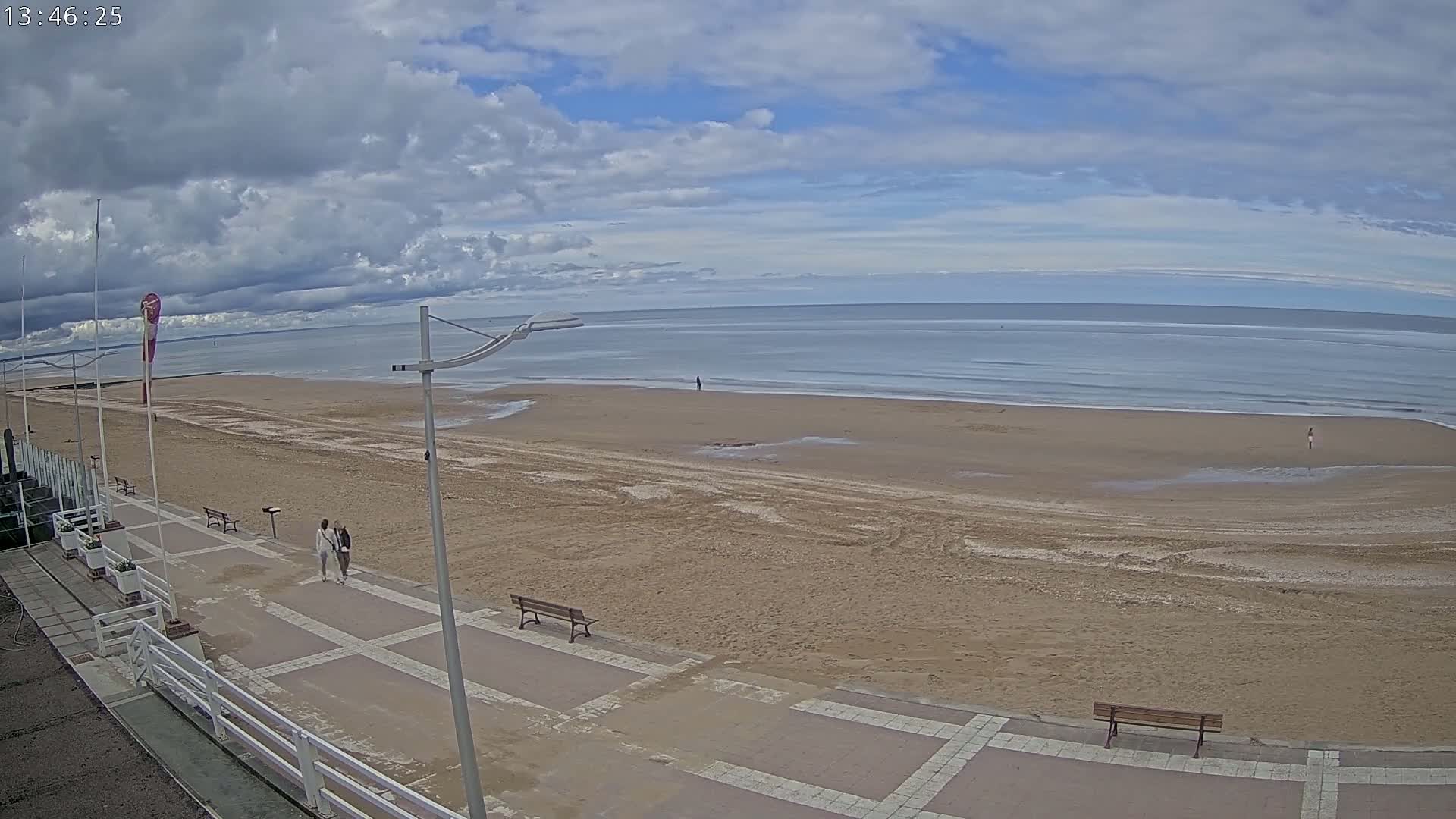 A mostly sunny day shows a wide sandy beach with a few people and benches along a paved walkway, with the calm ocean in the background.