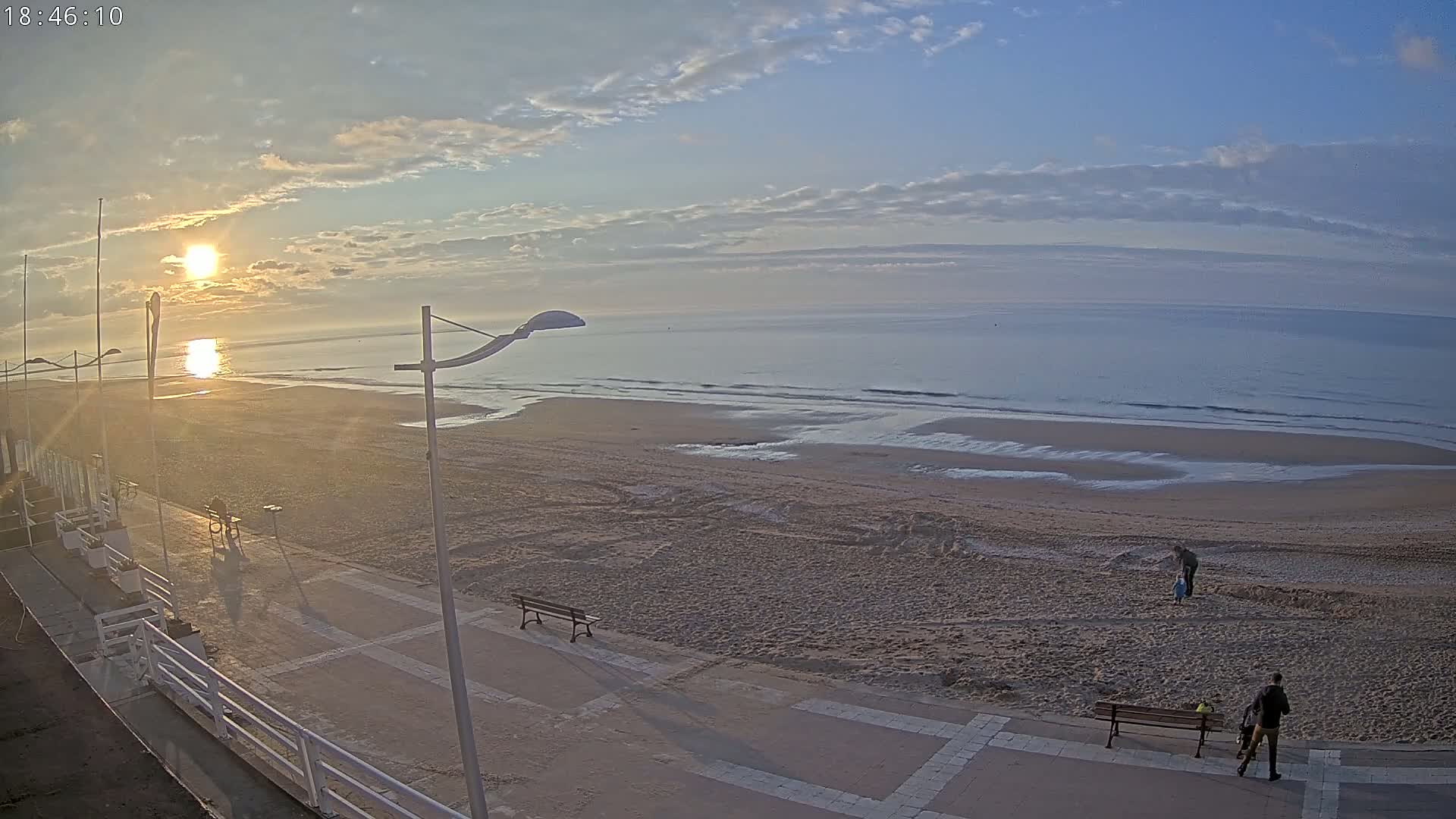 A sunrise illuminates a mostly empty sandy beach with calm waves, a boardwalk, and several people walking near benches.