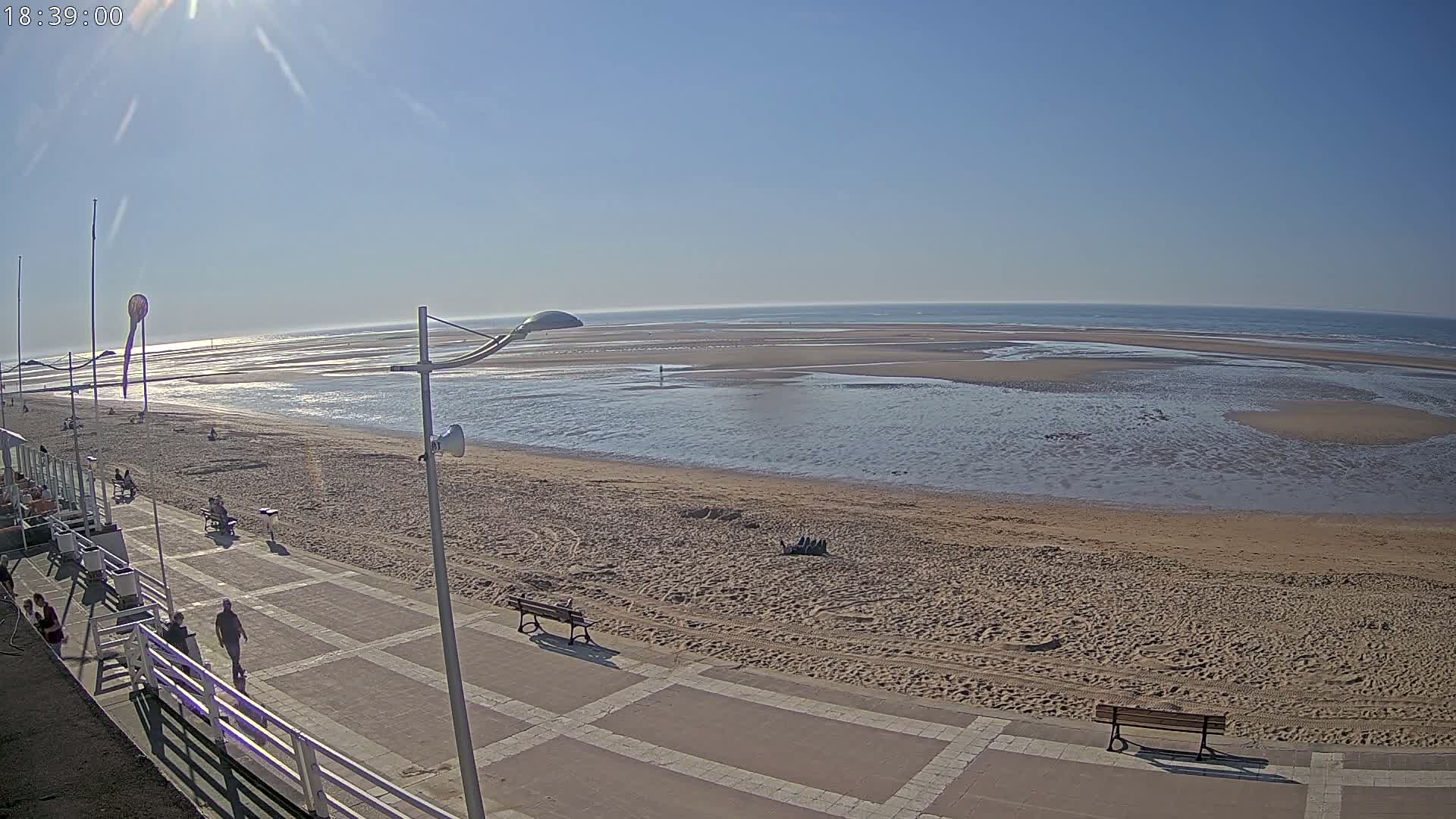 A mostly sunny day at a beach shows a sandy shore, calm water receding at low tide, and a paved walkway with several people and benches.