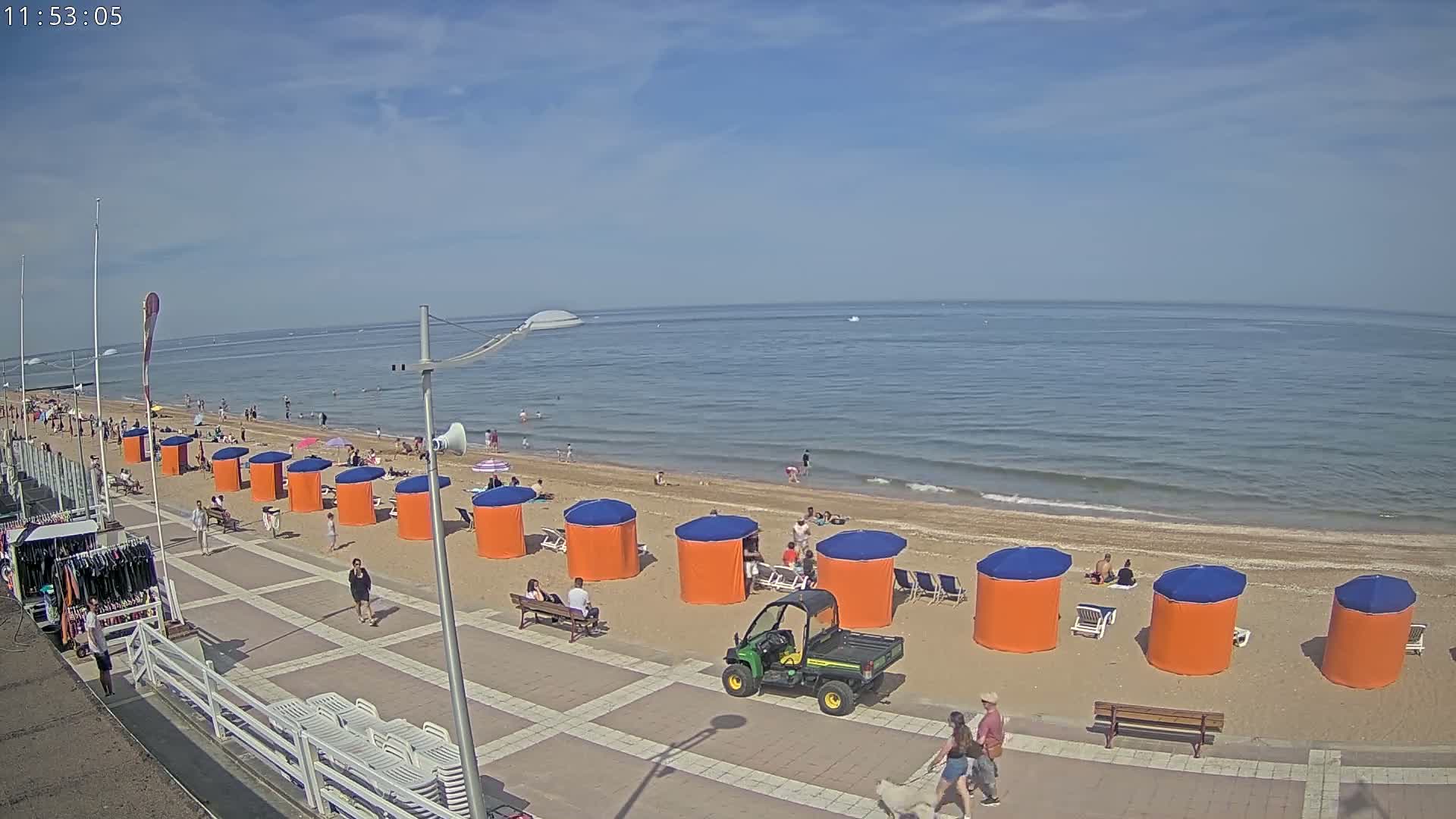 A sandy beach with orange and blue beach umbrellas, people swimming and sunbathing, and a utility vehicle is visible on a sunny day.