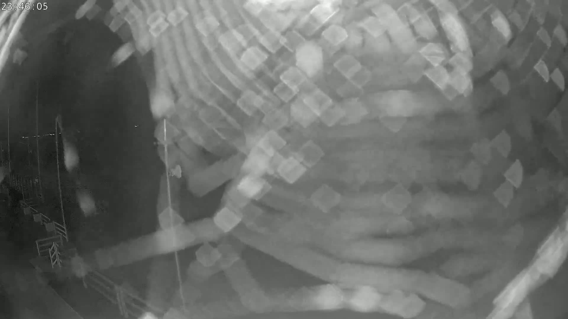A blurry, grainy, nighttime security camera image shows an indistinct object or scene with faint outlines of what may be structures or vehicles.
