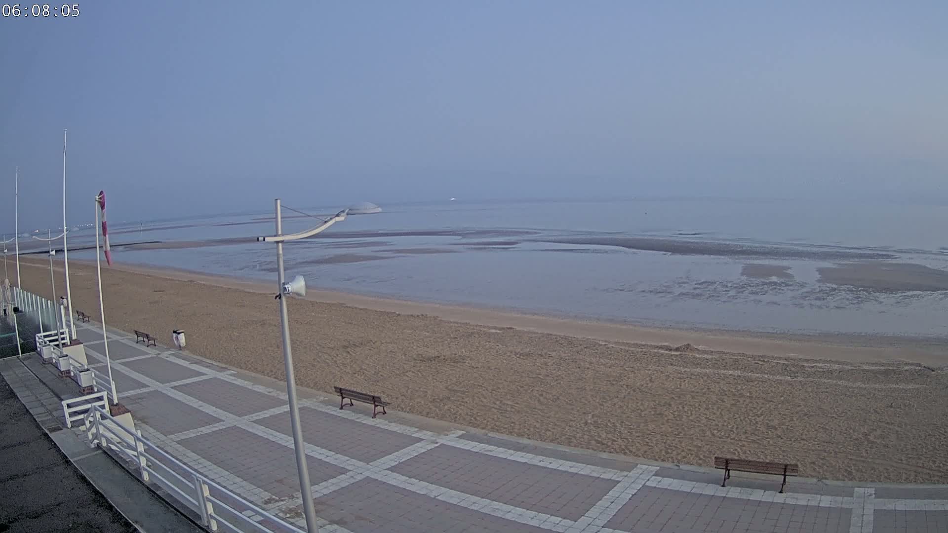 A sandy beach with a paved walkway and benches is visible, overlooking a calm, hazy ocean at low tide under a clear, pale sky.