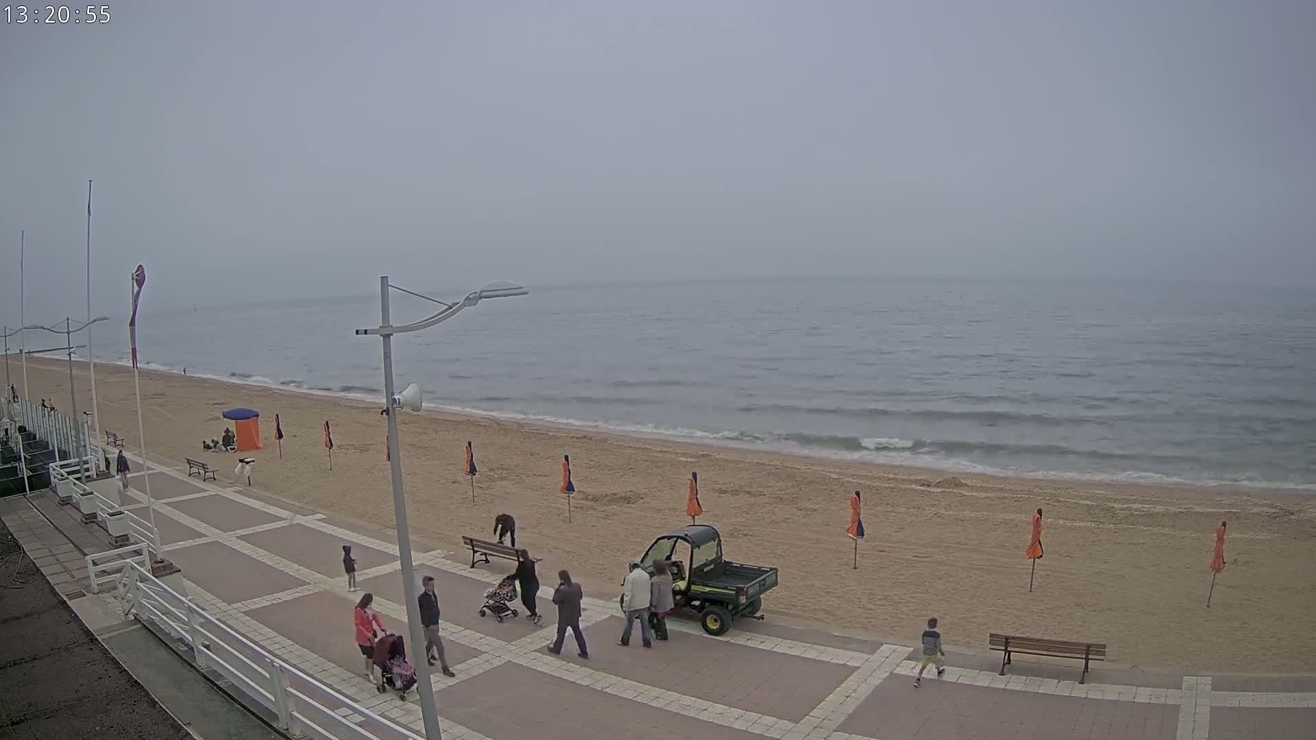 A mostly overcast day at a beach shows a sandy shore with several people walking along a paved boardwalk, some beach umbrellas, and a small utility vehicle.
