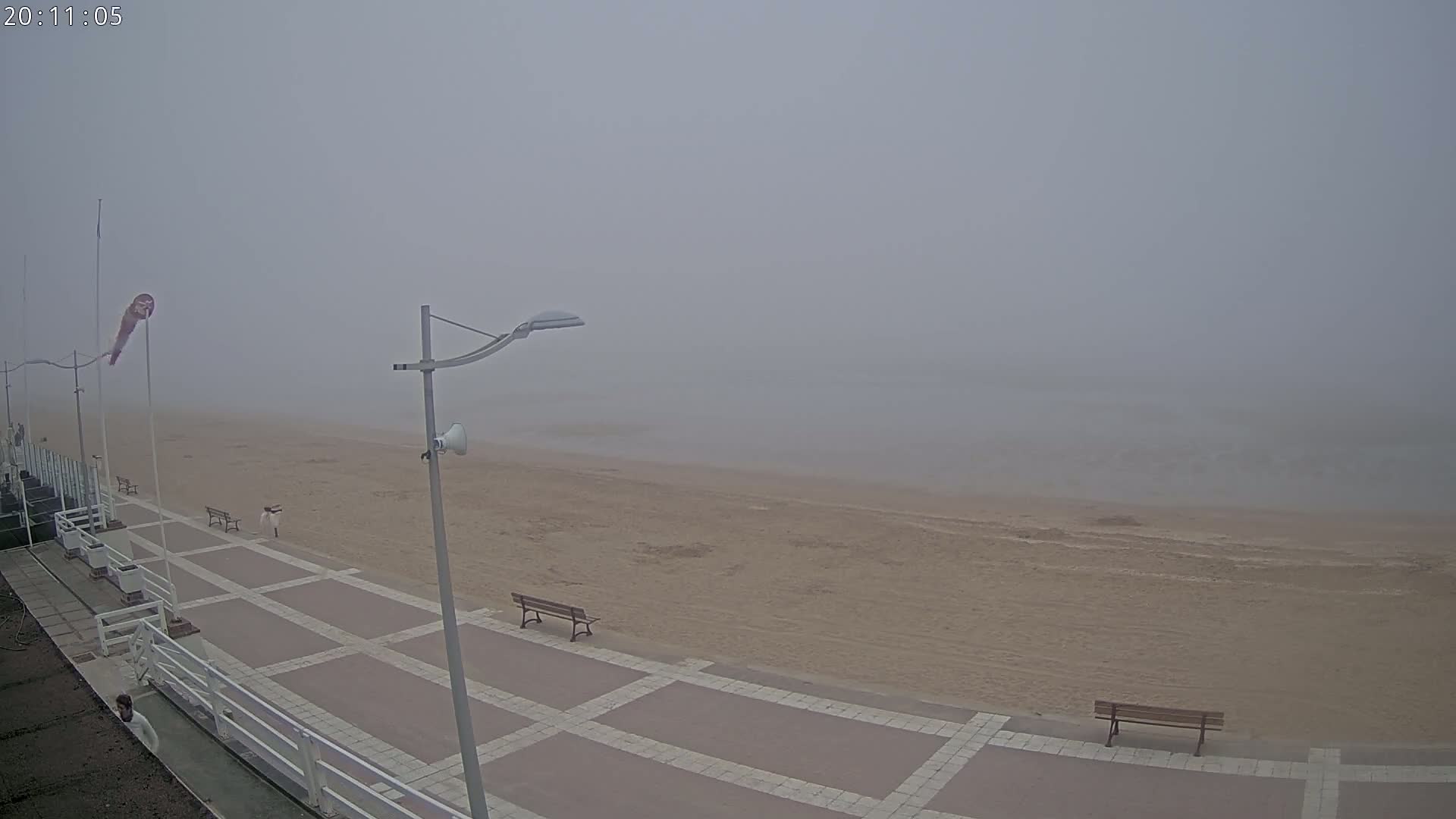 A foggy beach scene shows a paved walkway with benches overlooking a sandy beach and calm water.
