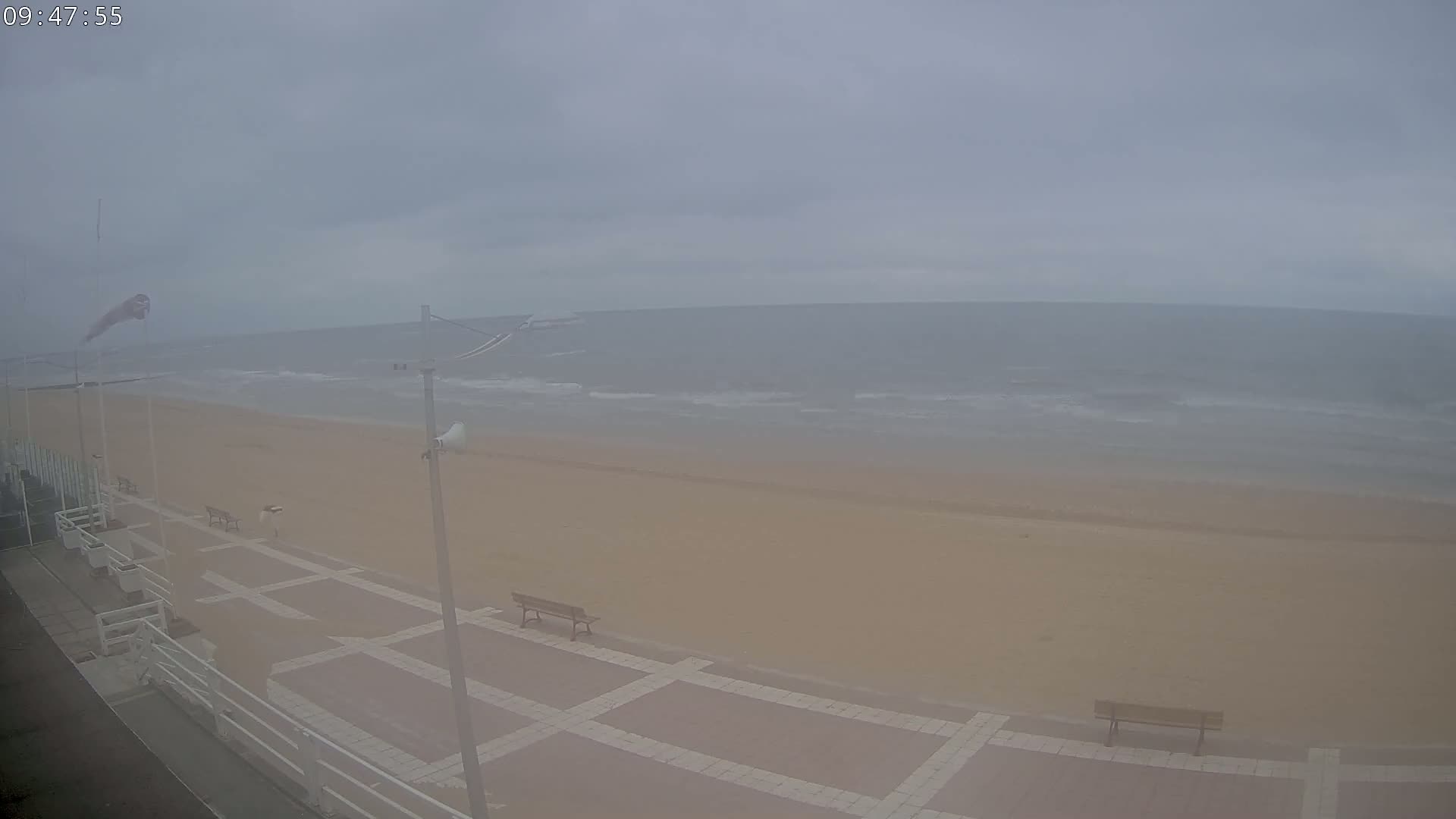 A hazy, overcast day reveals a sandy beach with a paved walkway, benches, and a calm ocean in the distance.
