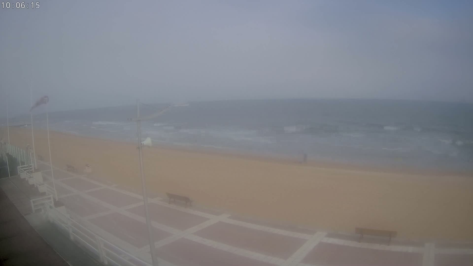 A hazy, overcast day shows a sandy beach with small waves, a boardwalk with benches, and a few distant figures.