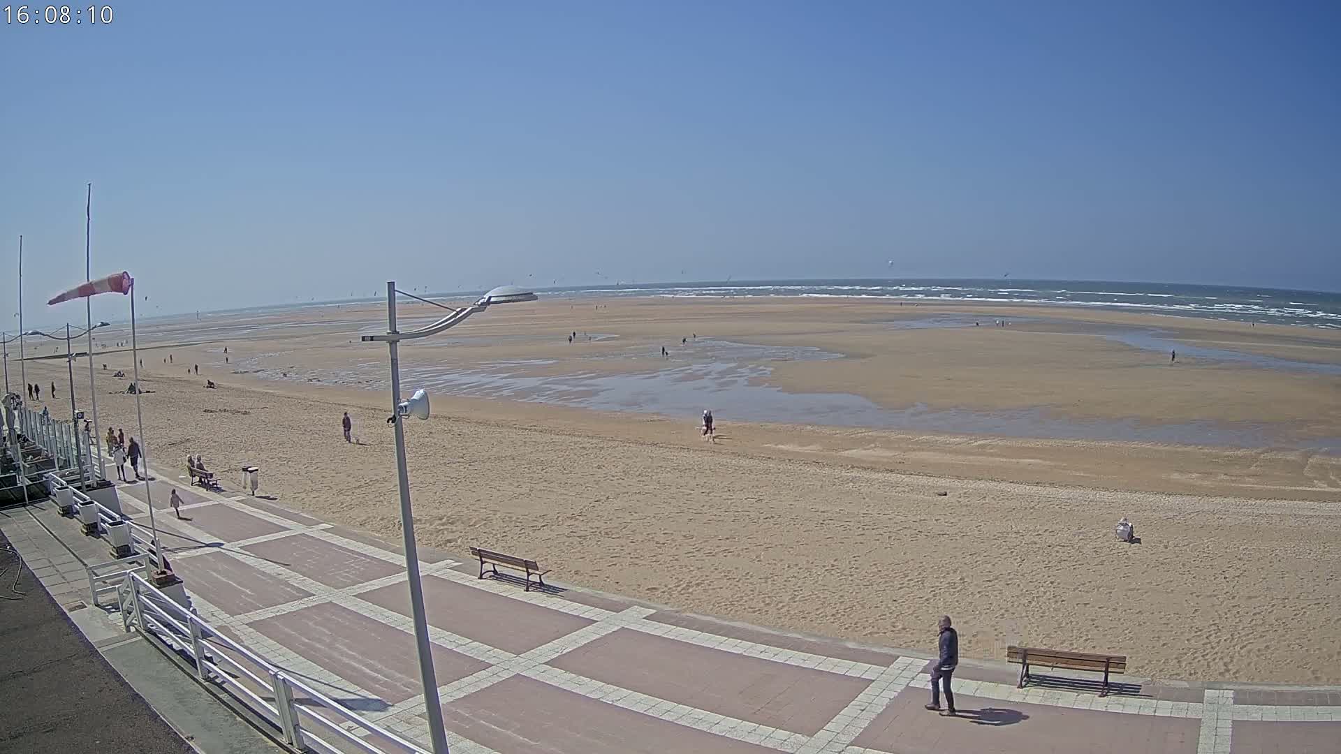 A wide sandy beach with a few people and benches on a paved walkway, under a clear blue sky.