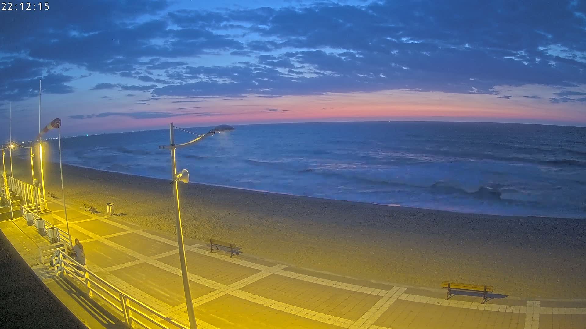 A mostly-clear twilight sky over a dark-blue ocean with small waves lapping a sandy beach, where a paved walkway with benches is illuminated at night.