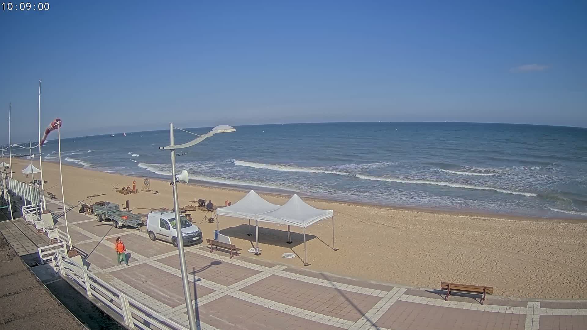 A sandy beach with gentle waves under a clear blue sky, featuring a paved walkway with several people, a white van, and two pop-up tents.