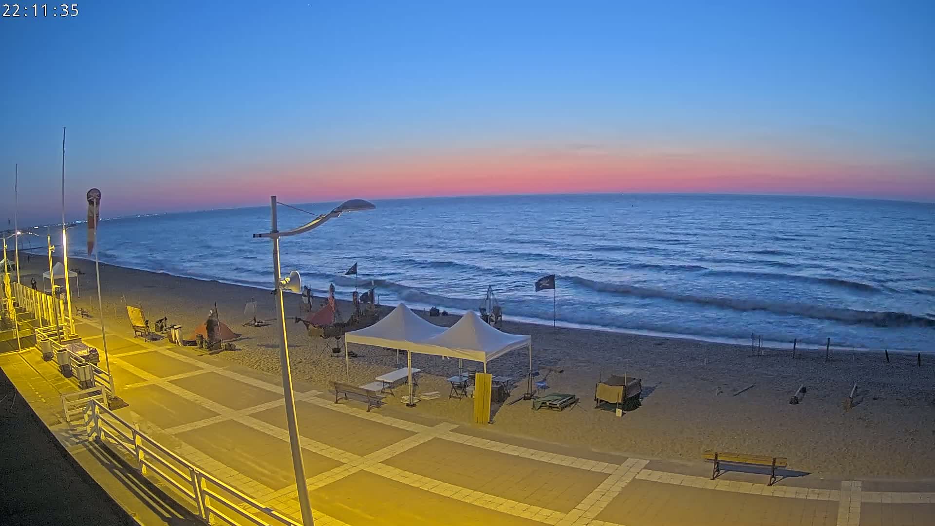 A mostly empty beach at twilight, with a paved walkway, several tents, and some equipment set up on the sand, under a pink and blue sky.