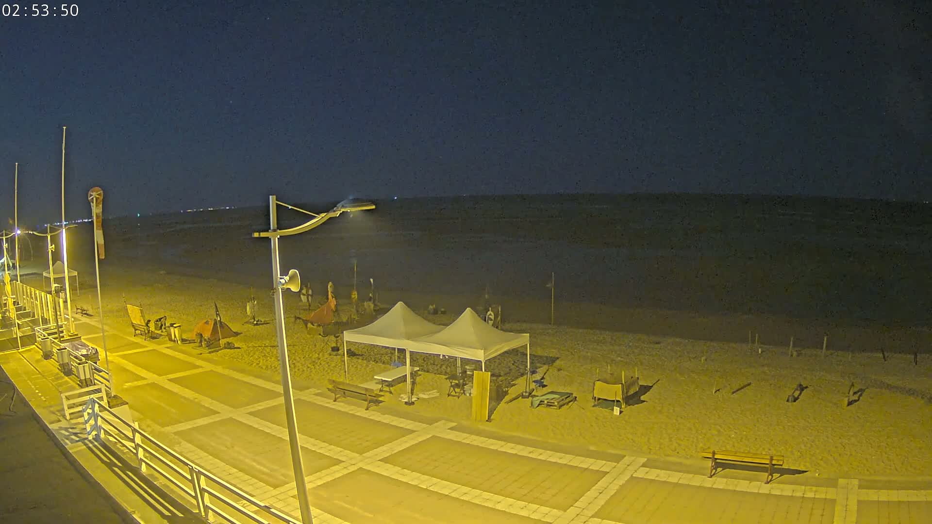A nighttime beach scene shows a sandy shore with several tents and equipment set up near a paved walkway and boardwalk under a dark, clear sky.