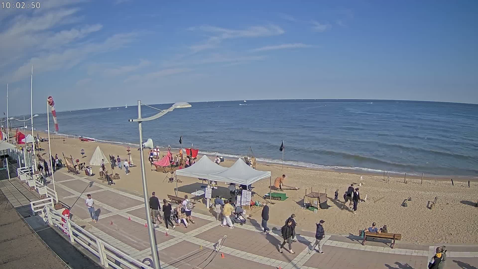A sunny day at a beach shows people gathered around various structures on the sand and boardwalk, with a calm ocean in the background.