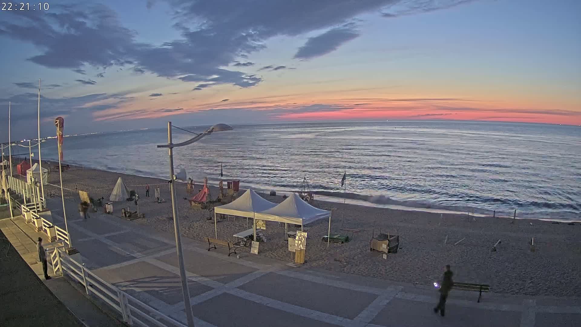 A mostly clear sunset over a beach with several small tents and a few people walking along a paved walkway.