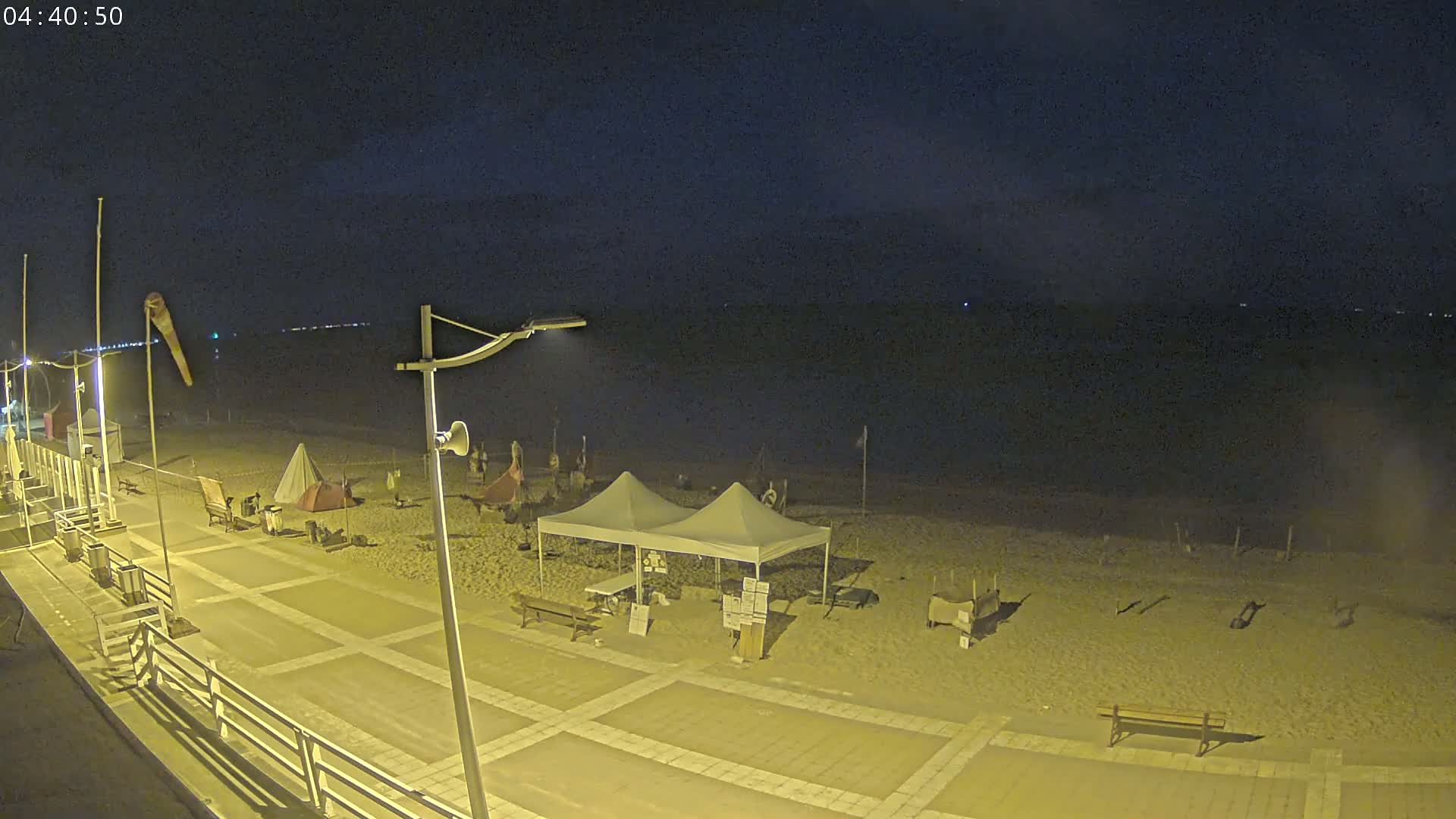 A nighttime view of a sandy beach with several tents and beach equipment, a boardwalk, and streetlights illuminating the scene under a dark sky.