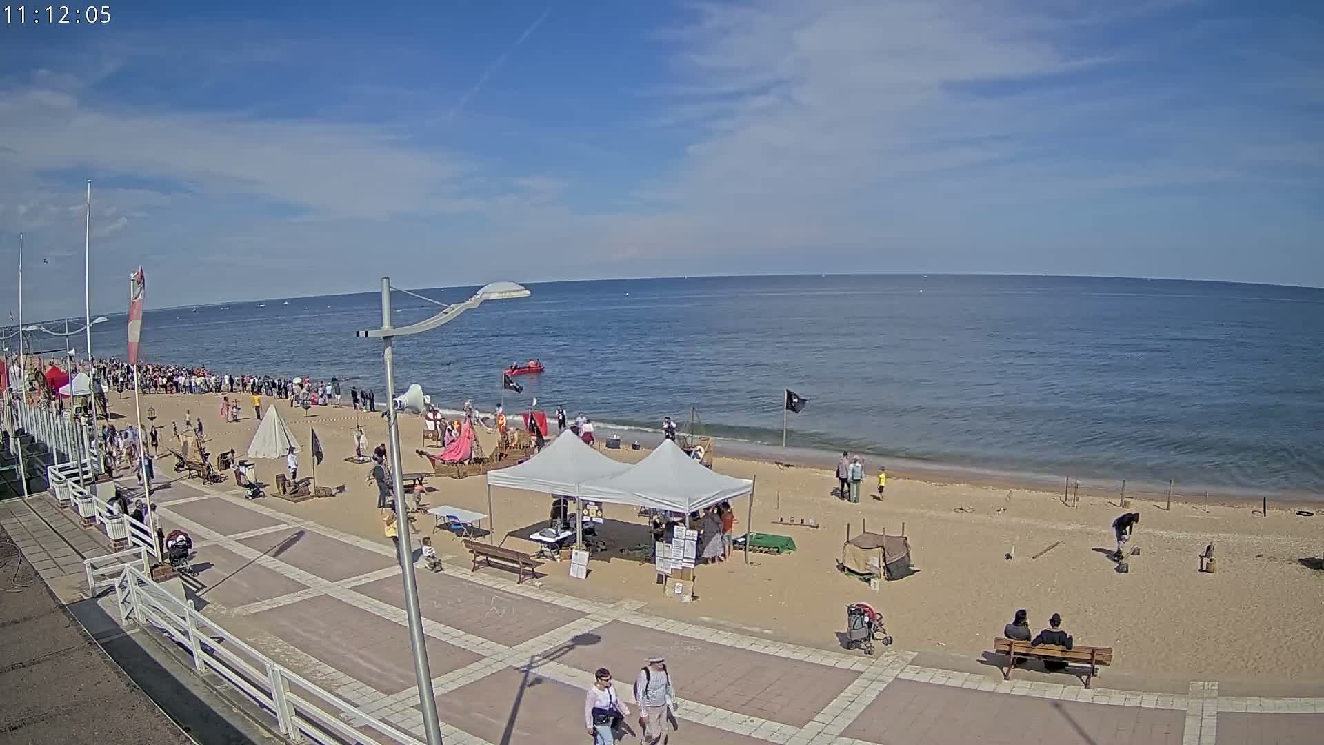A sandy beach with a large crowd of people, several tents, and small boats in the ocean under a sunny sky.