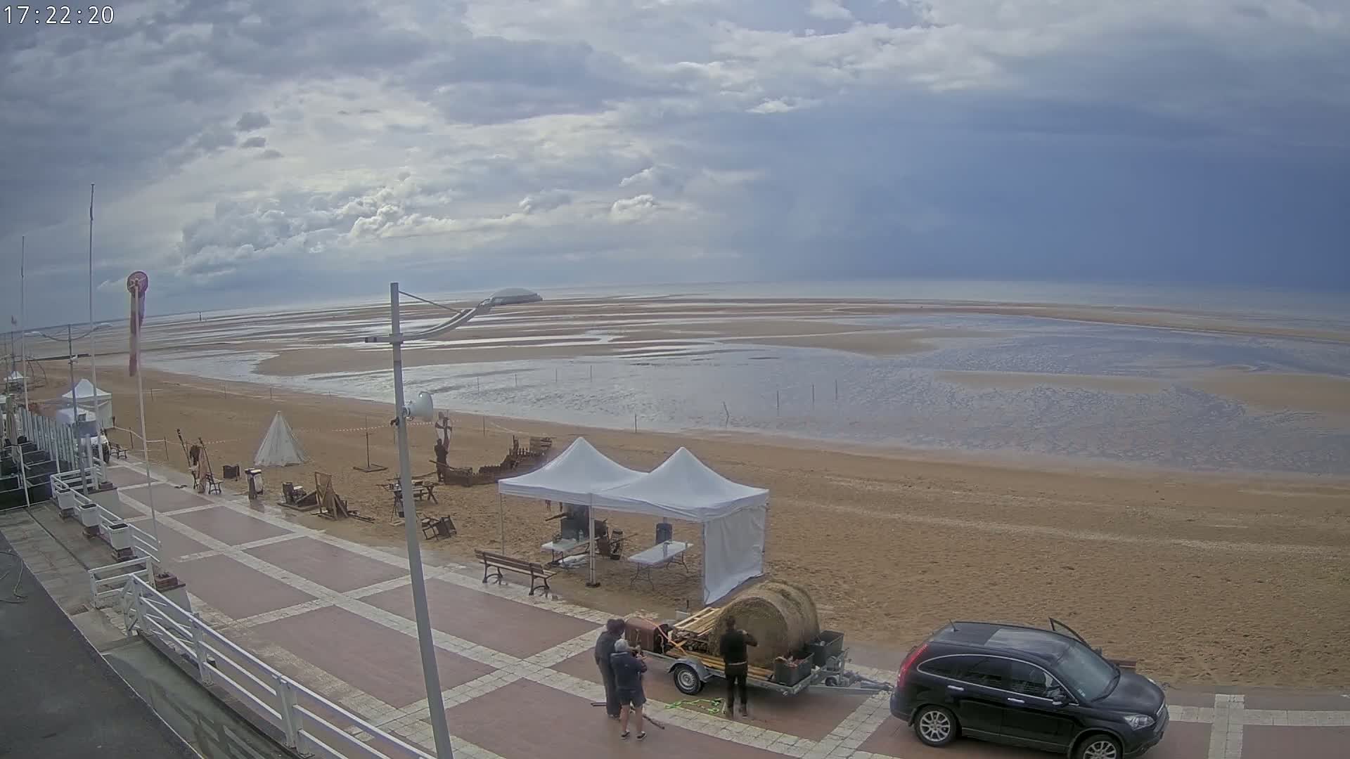A sandy beach with calm water under a cloudy sky, features a paved walkway, several people near a trailer loaded with hay bales, and various items set up on the sand near two white tents.