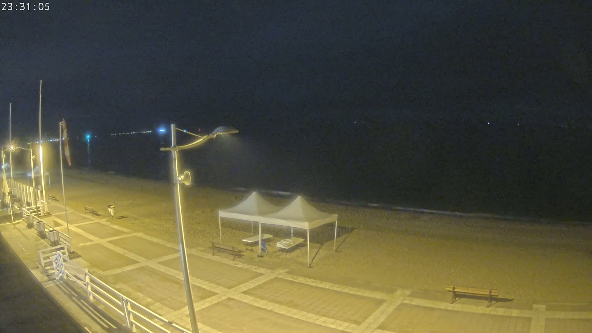A nighttime beach scene shows a sandy shore with a white pop-up tent and several benches under illuminated streetlights, next to a paved walkway and a calm dark sea in the background.