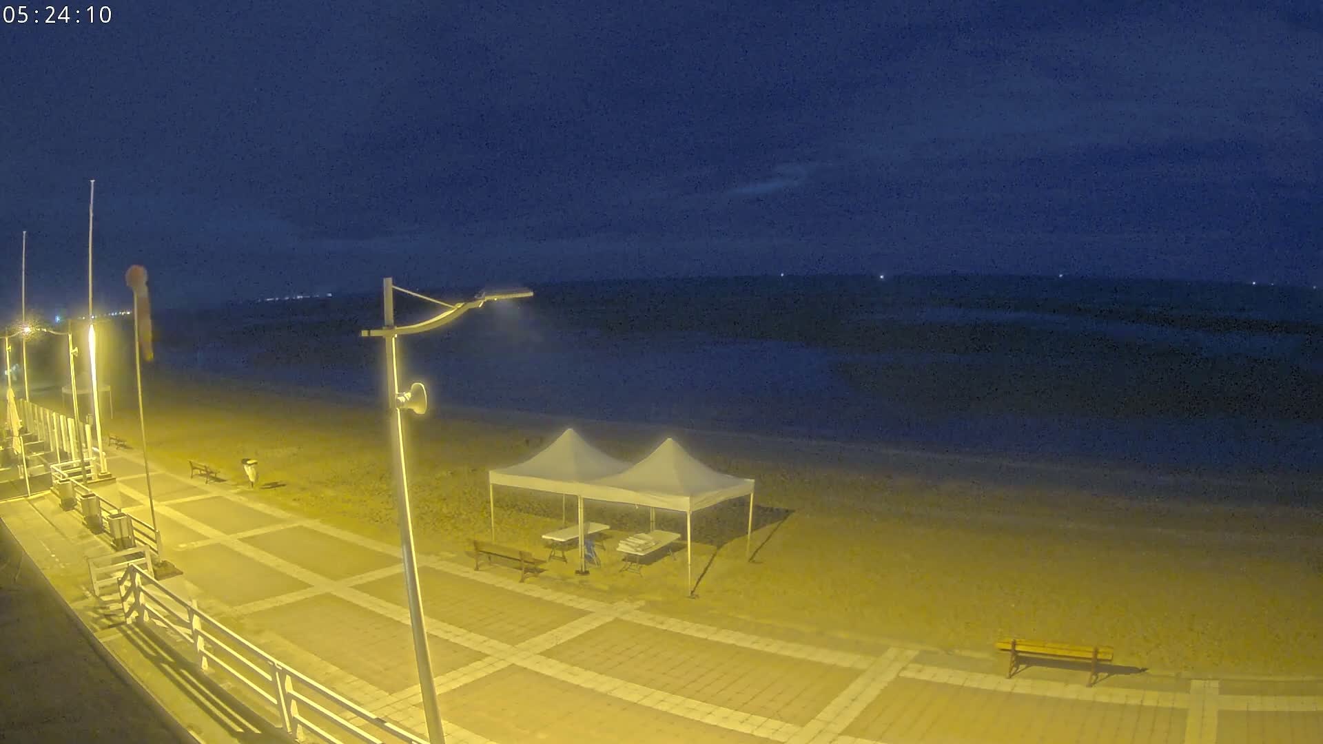 A sandy beach at night, illuminated by artificial lights, features a couple of pop-up tents and a boardwalk.
