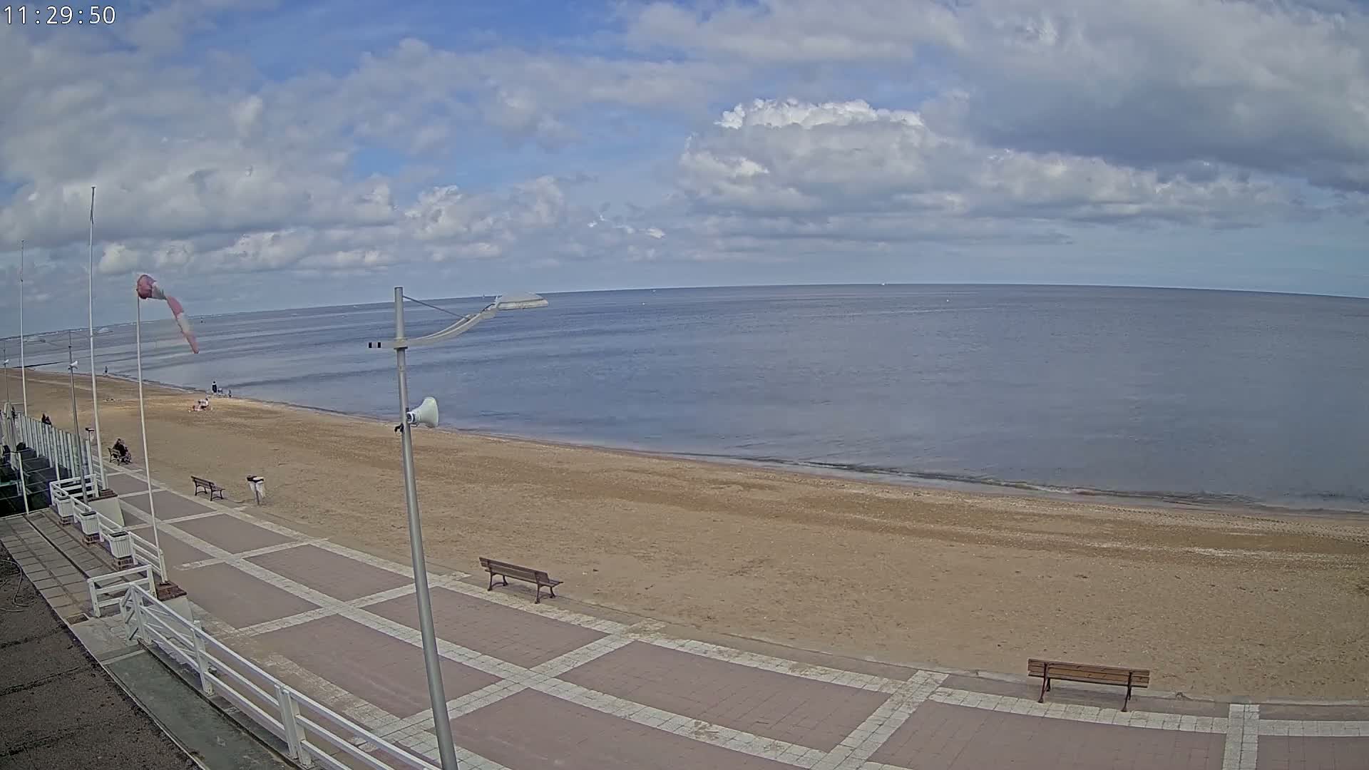 A sandy beach with a paved walkway and benches stretches alongside a calm sea under a partly cloudy sky.