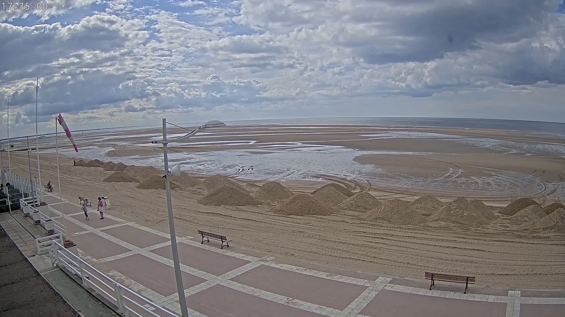 A mostly cloudy day reveals a wide sandy beach with piles of sand along a paved walkway, a few people strolling, and a calm ocean in the distance.