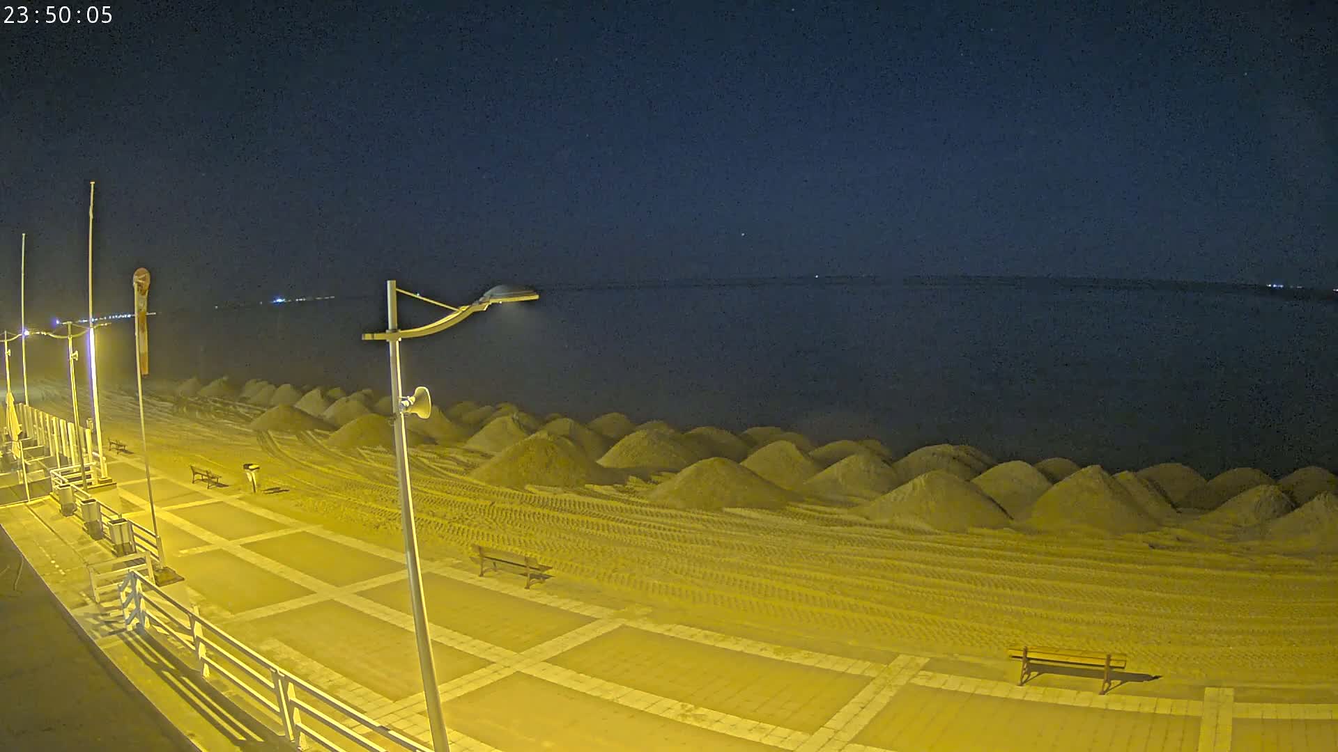 A nighttime view of a beach boardwalk illuminated by streetlights, showing a stretch of beach with numerous small piles of sand next to the water under a clear, dark sky.