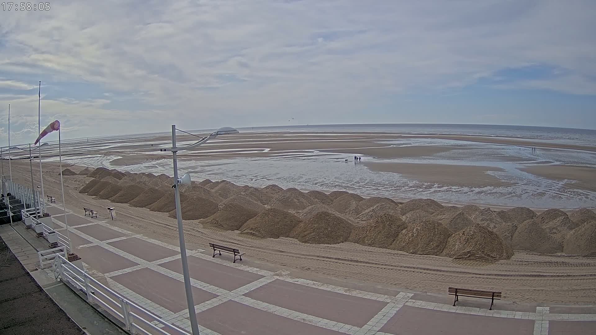 A mostly cloudy day reveals a wide sandy beach with a row of sand piles near a paved walkway, and a few people walking on the wet sand near the water.