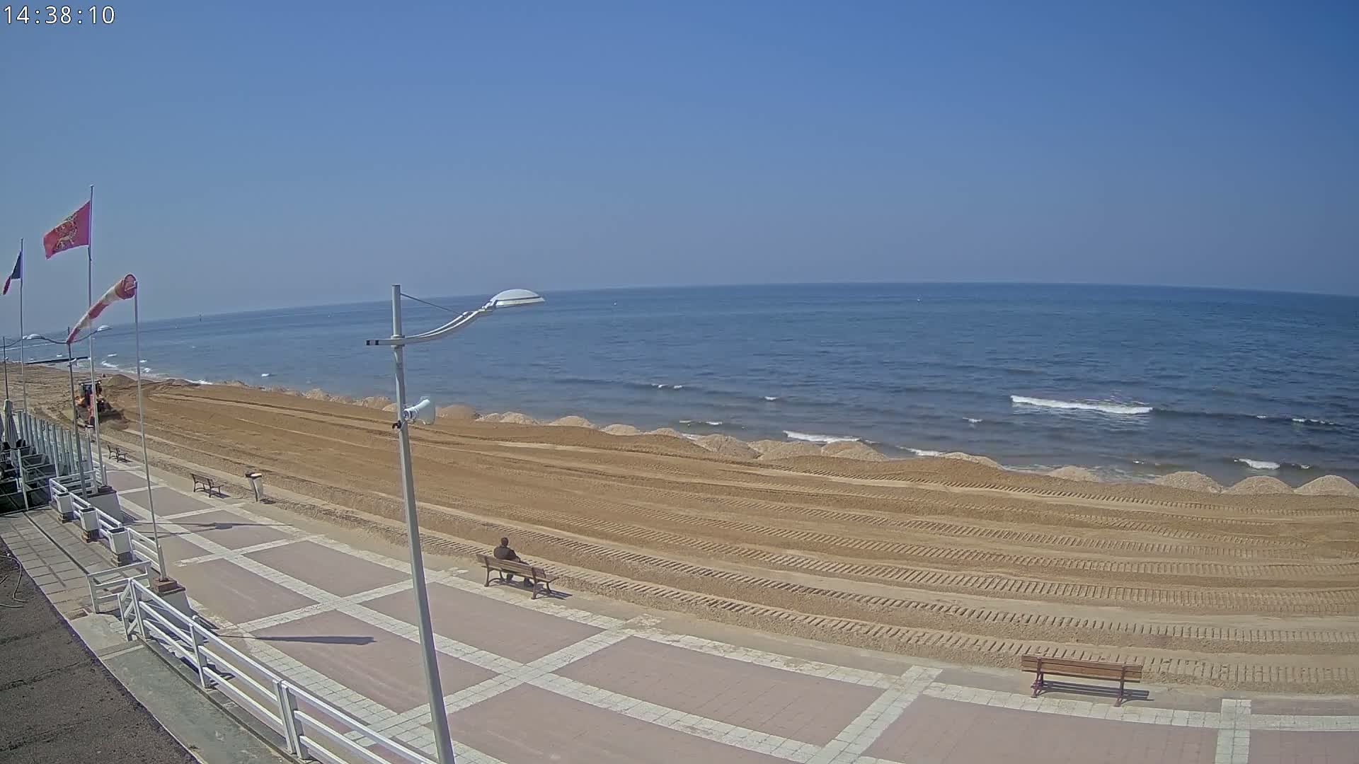 A sandy beach with a person sitting on a bench, under a clear blue sky, is being worked on by machinery near a paved walkway.