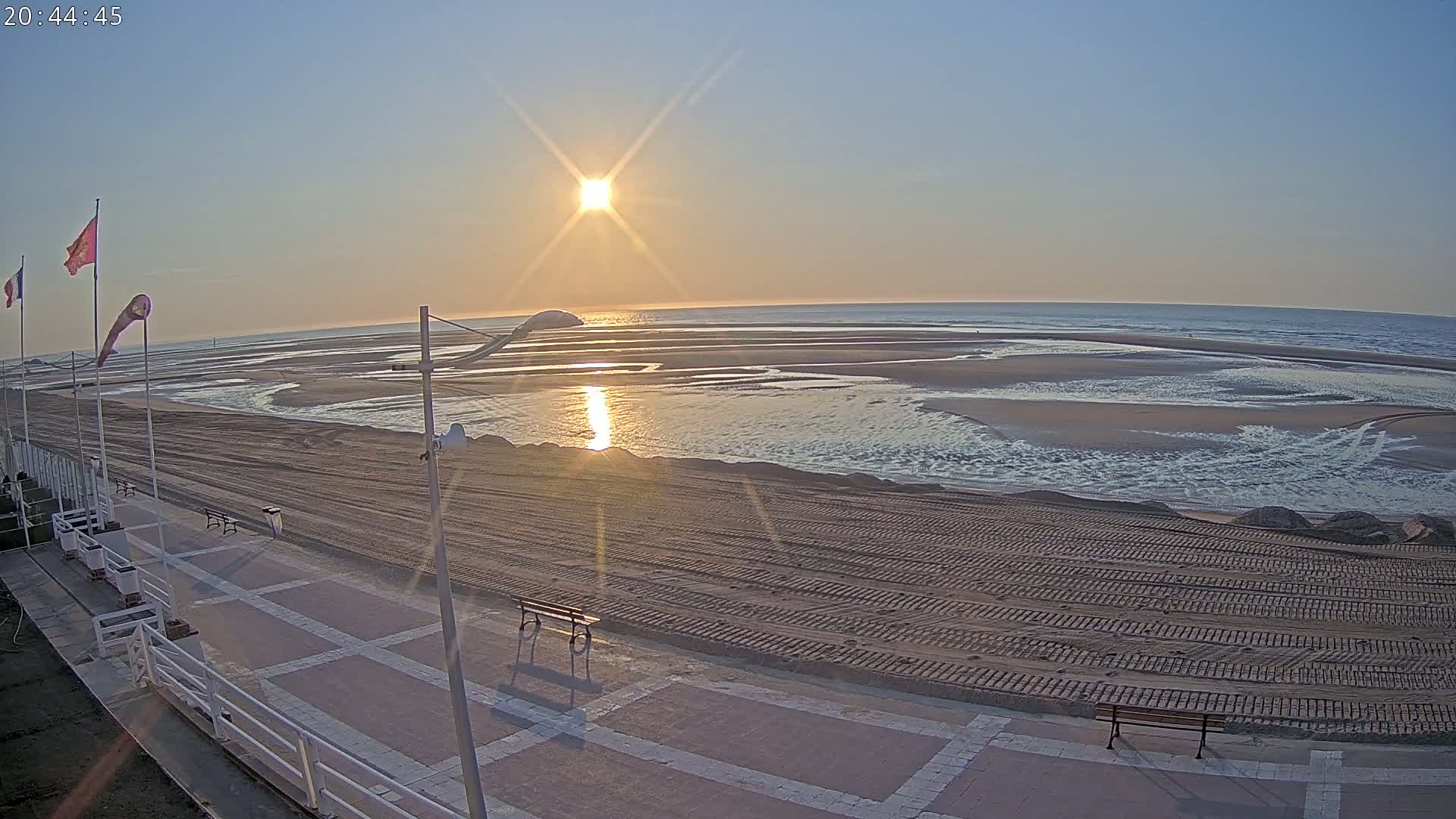 A sandy beach at sunrise, with a paved walkway and benches, shows the ocean receding to reveal tidal pools and channels.
