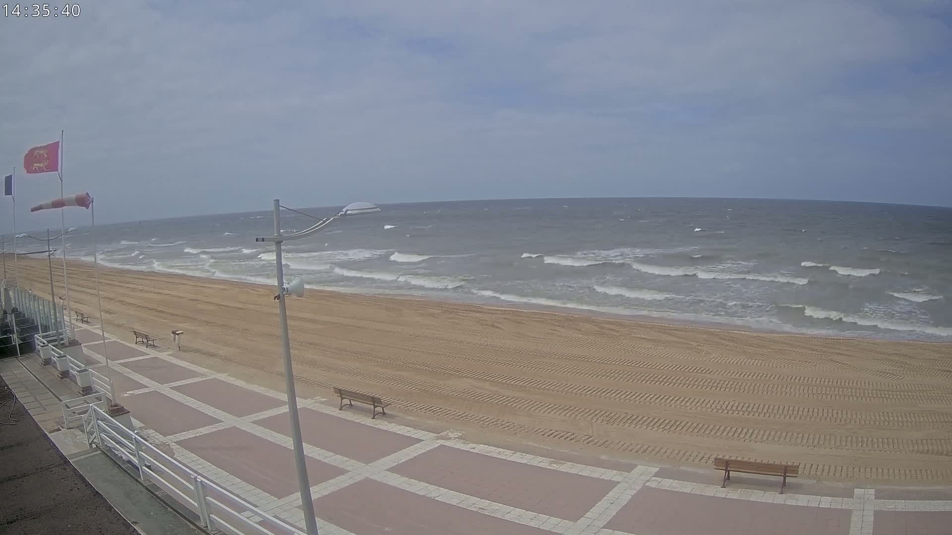A sandy beach with a paved walkway and benches is shown, overlooking a wavy ocean under a mostly cloudy sky.