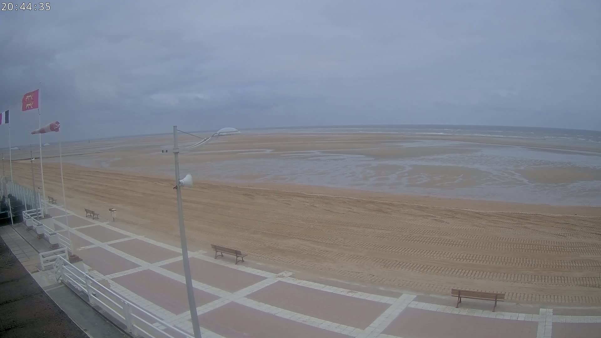 A wide, sandy beach stretches to a calm, grey sea under an overcast sky, with a paved walkway and benches visible in the foreground.