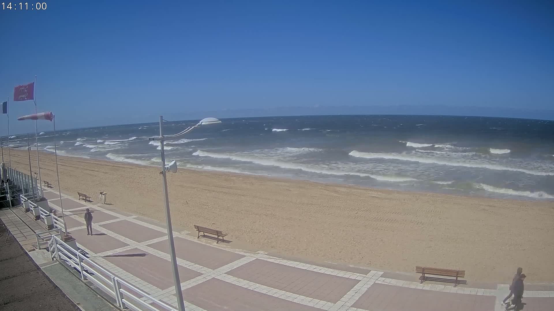 A sandy beach with a paved walkway and benches is shown, under a clear blue sky; moderate waves are visible in the ocean.