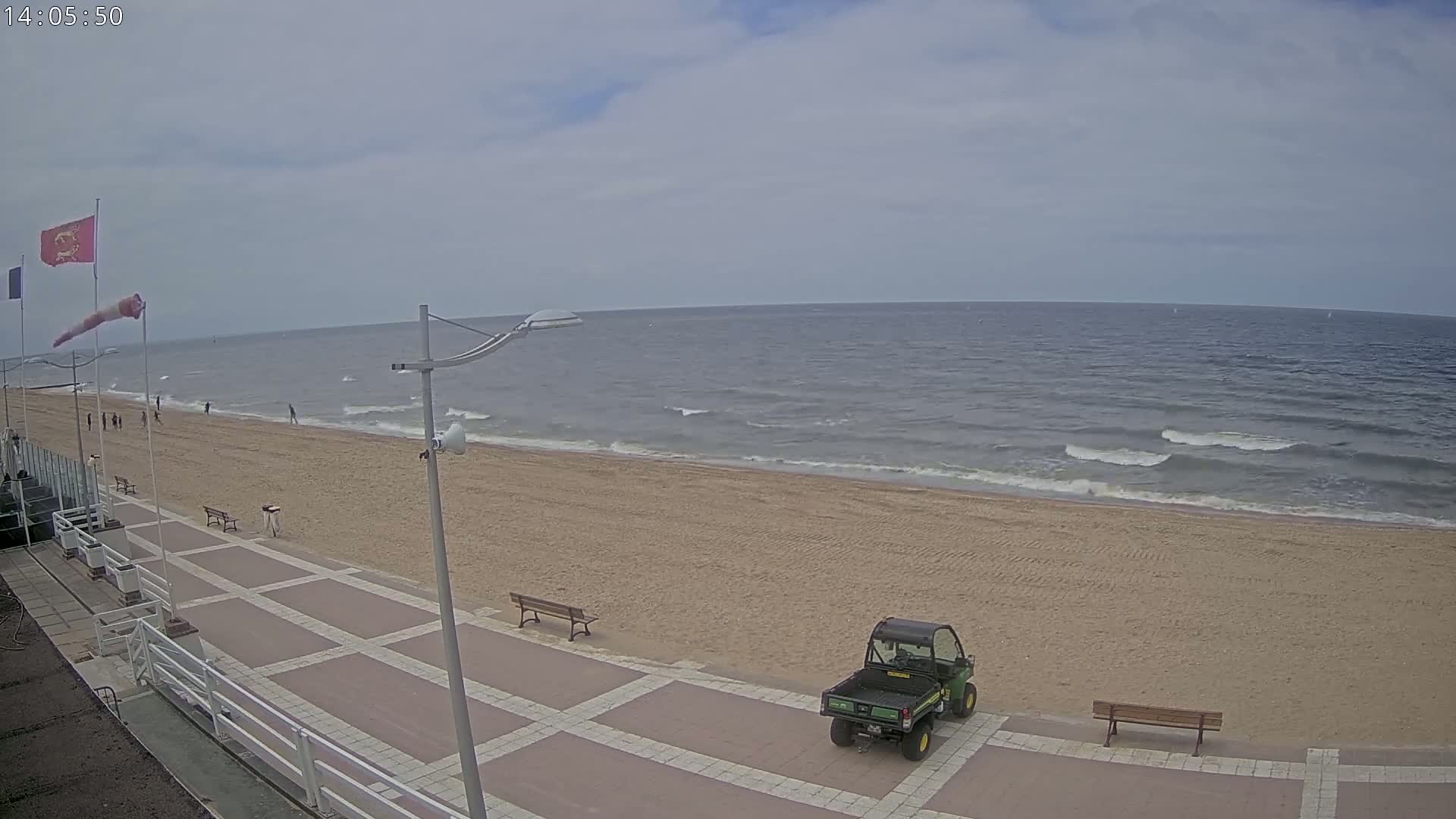 A sandy beach with a paved walkway, a few benches, a small utility vehicle, and a calm sea under a cloudy sky.