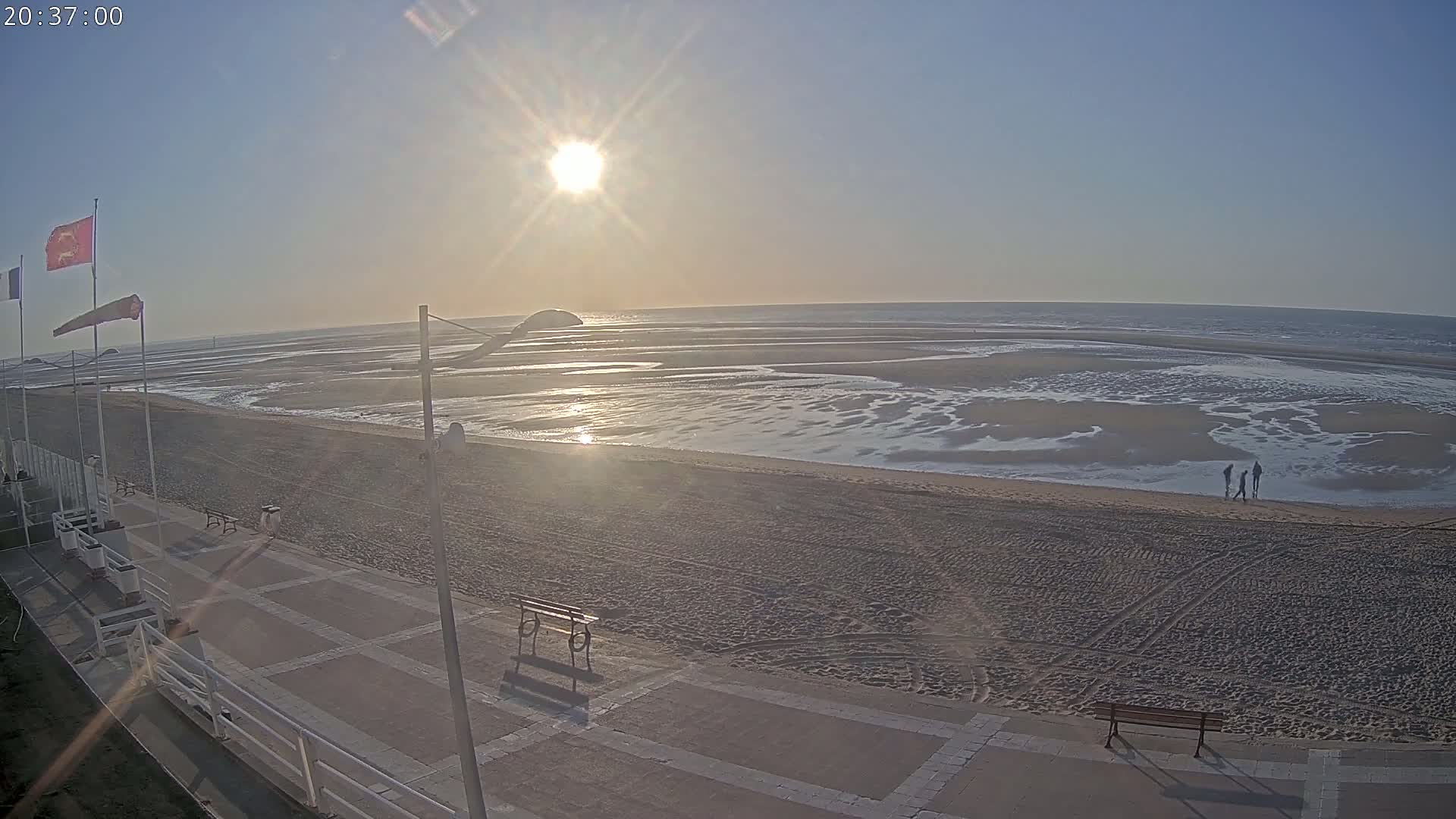 A sandy beach with a paved walkway and a few people walking near the water at sunset under a clear sky.